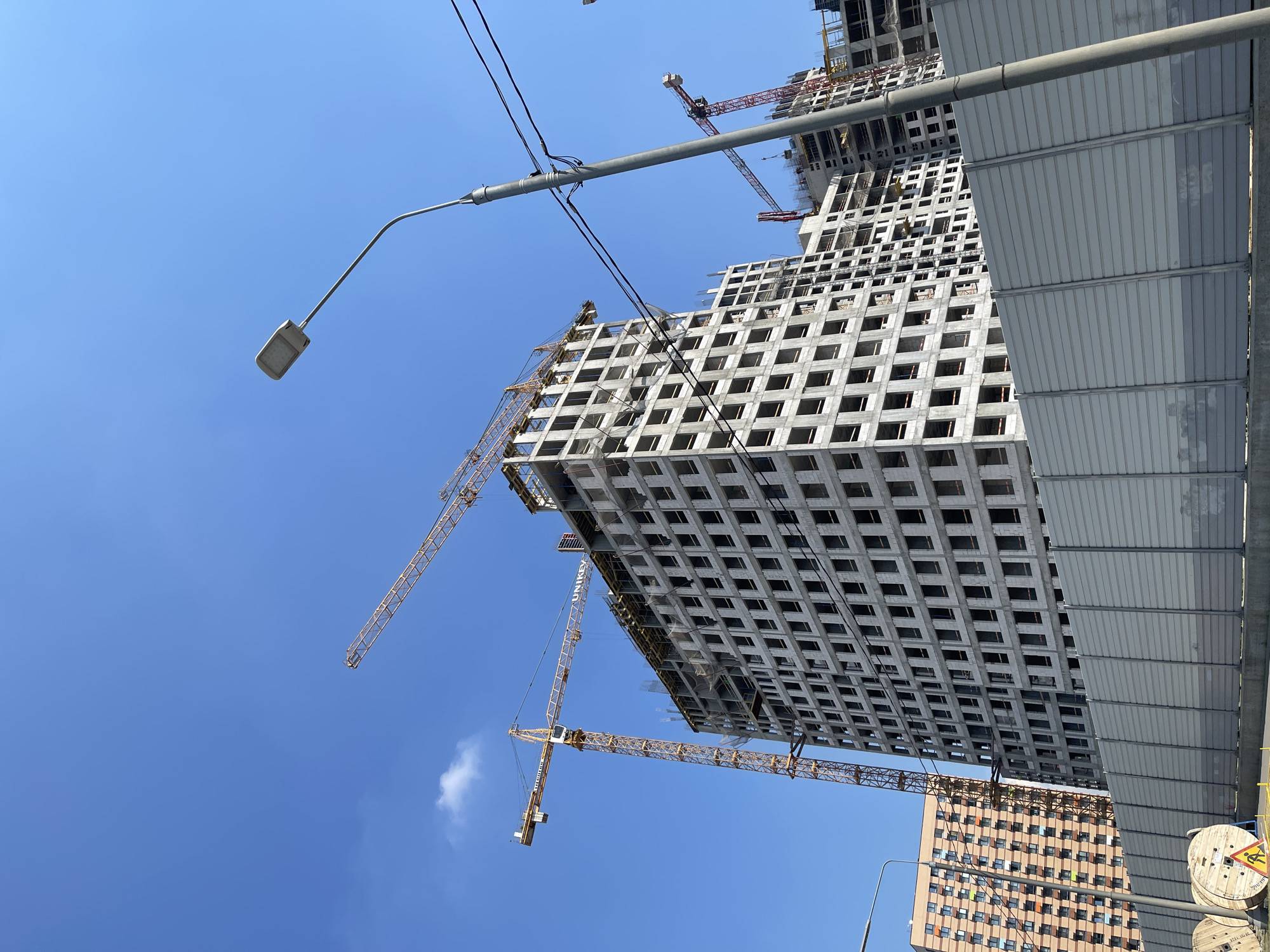 In Kommunarka, Russia, a construction site is depicted under a clear blue sky. Several multi-story buildings are in various stages of development. A large, unfinished concrete building with numerous window openings dominates the center of the frame, with multiple yellow tower cranes positioned around it. To the left, another residential building with a patterned facade is visible. A grey, corrugated metal barrier runs along the bottom of the image, separating the construction area from the foreground. A street lamp and power lines are also present. In the lower left corner, two large wooden spools, likely for cables, are situated on the ground, one with a yellow construction safety sign attached.