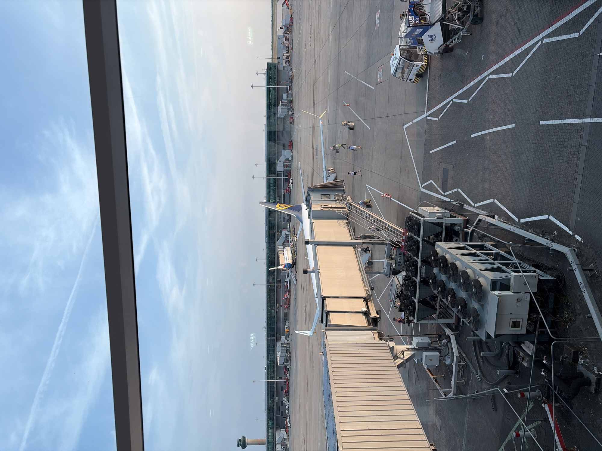 This image shows an aerial view of an airport tarmac, taken from inside a building, likely a terminal, looking out through a window. The scene is set on a sunny day with a clear blue sky dotted with wispy clouds. In the foreground, a jet bridge extends towards an airplane. The airplane has "RYANAIR" written on its tail fin. Several people can be seen on the tarmac, some walking near the airplane and others standing by vehicles.
Various airport vehicles and equipment are visible on the tarmac, including what appears to be a baggage tug or service vehicle with "BLUE HANDLING" printed on its side and the number "233". Further down the tarmac, more aircraft and airport infrastructure, including terminal buildings with large windows, are visible. A control tower can be seen in the distance on the left side of the image. The ground is paved with cobblestones in some areas and asphalt in others, marked with white lines indicating taxiways and parking spots. The overall atmosphere appears calm and functional, typical of an active airport environment. The time of day seems to be daytime, given the bright sunlight and clear sky. The location is identified as Uttlesford, United Kingdom.