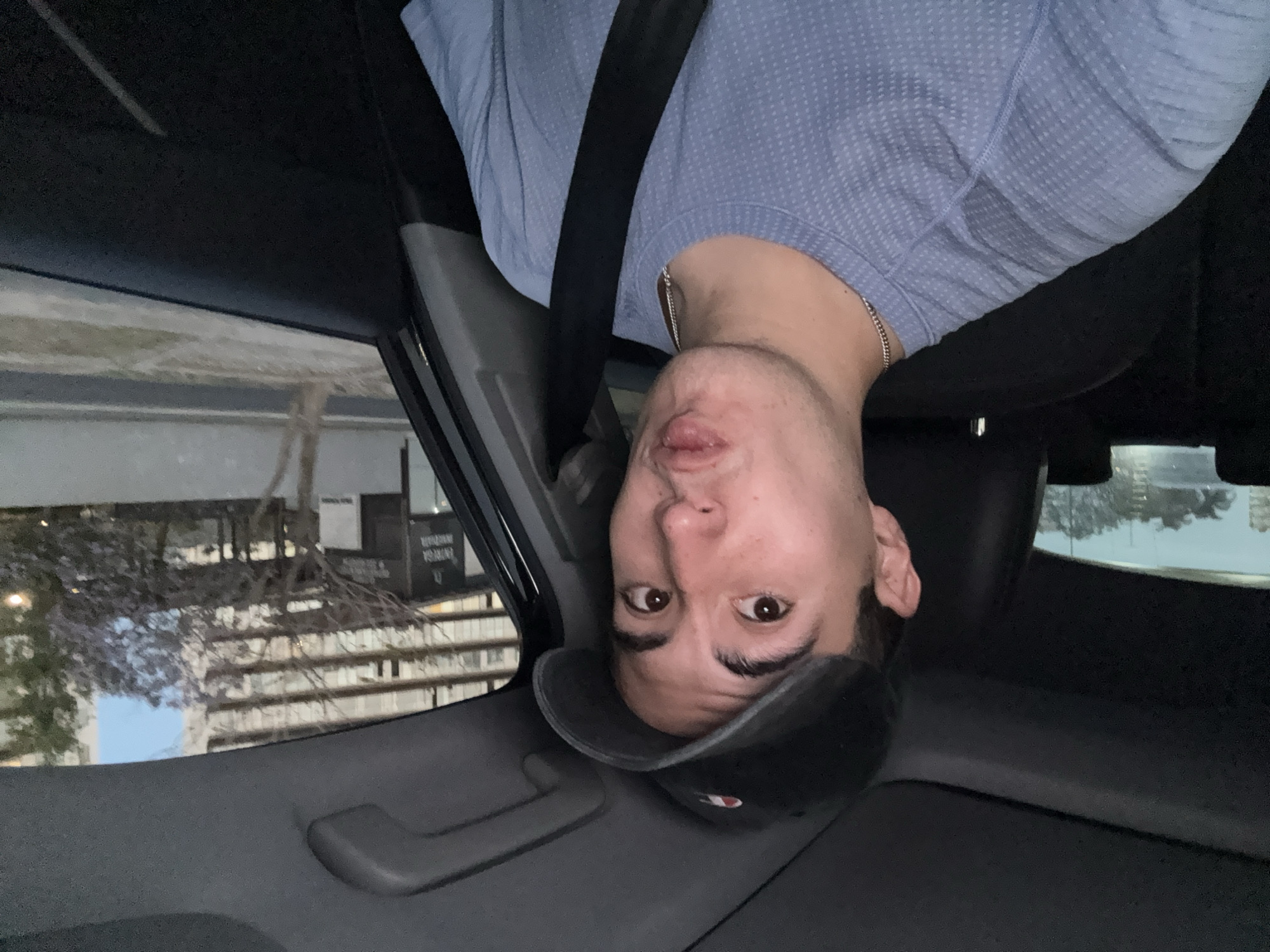 A portrait-oriented image captures a man's upper body and face, inverted, inside the passenger cabin of a vehicle. The man wears a light blue, textured short-sleeved shirt, a dark baseball cap, and a silver chain around his neck. He is secured by a dark seatbelt diagonally across his chest. His face, positioned towards the bottom-center of the frame, displays a playful expression with puckered lips and wide, brown eyes looking directly at the viewer.
The interior of the car is visible along the bottom and right edges of the frame, showing a gray headliner and a grab handle. Through the car window, visible on the left side of the frame (when the man is inverted), an outdoor urban or suburban scene is captured, also appearing inverted. This background features multi-story buildings with numerous windows and balconies, along with trees, some bearing what appear to be purple blossoms. Patches of light sky are also visible.
On a dark, inverted sign seen through the window, white text is partially discernible, appearing to read "ENERGÍA RENOVABLE". The lighting suggests it is daytime, possibly late afternoon or early evening. The overall impression is one of humor or lightheartedness due to the man's expression.