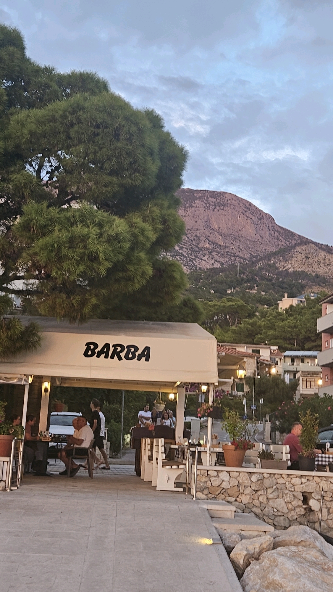The image depicts an outdoor restaurant scene in Općina Tučepi, Croatia, at dusk. A white awning with the black lettering "BARBA" is prominently displayed above the entrance to the establishment. The restaurant features outdoor seating with benches and tables, some of which are set with checkered tablecloths. Several patrons are seated at tables, engaged in dining and conversation.
To the left of the restaurant, a large, mature pine tree with dense green foliage dominates the foreground. In the background, a rugged, rocky mountain rises above a hillside covered in greenery and scattered buildings, indicating a coastal or mountainous region. The sky is overcast with soft clouds, suggesting the late hour of the day. The ground is paved with light-colored stones, and a low stone wall with potted plants borders a section of the seating area. The overall ambiance is relaxed and communal, characteristic of a European seaside town.