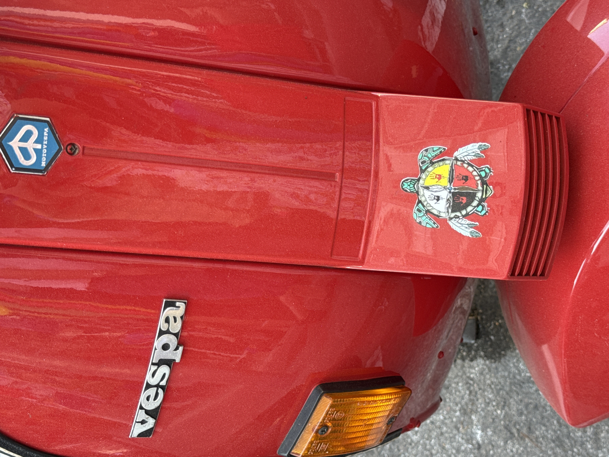 The image shows a close-up of the rear of a red Vespa scooter parked on a gray asphalt surface. The scooter has a silver "Vespa" logo on its left side. Above the logo, on the scooter's body, is a hexagonal blue and white "MOTO VESPA" emblem. Below the emblem and centered on the scooter's rear panel is a sticker of a turtle. The turtle sticker has a circular design on its shell divided into four quadrants, each containing a handprint and a different color: black, yellow, red, and white. The turtle has a turquoise shell outline and light blue feathers on either side. The scooter has a clear orange turn signal on its left side. The scene is static, with no people or actions depicted. The environment appears to be an outdoor urban setting.