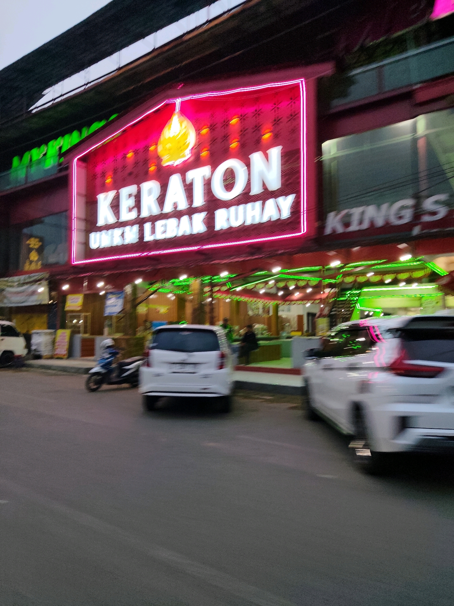 The image captures a street scene in Rangkasbitung, Indonesia, focusing on a building with a prominent neon sign that reads "KERATON UNKM LEBAK RUHAY." The sign is illuminated with bright pink neon tubing surrounding white, block-style lettering. Above the main text, a graphic of a yellow, bulbous object, possibly a lamp or fruit, is depicted. To the right of the "KERATON" sign, another sign with the partial text "KING S" is visible.
The building itself appears to be a commercial establishment, with storefronts and possibly multiple levels. The facade features red and dark framing around the signs and windows. Green neon lights are strung along the lower level of the building, accentuating the entrance and covered walkways.
In the foreground, a street is visible with asphalt pavement. Several vehicles are present, including two white cars and a motorcycle. The motorcycle, ridden by a person wearing a helmet and a blue jacket, is moving towards the right side of the frame. The white cars are also on the road, with one partially obscuring the other. The scene suggests a bustling commercial area, likely in the evening or dusk, given the illuminated signs. There are also hints of people in the background, some possibly seated or standing near the building's entrance. The overall impression is of a vibrant urban environment with active commerce.