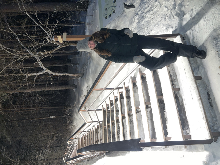 A female individual stands at the base of a long, illuminated, snow-covered wooden staircase in a dense pine forest, located within the Bobrovy Log Funpark area of Krasnoyarsk, Russia. The scene is depicted under low-light conditions, consistent with nighttime, with artificial lighting illuminating the staircase and immediate foreground.
The subject is a female with long, wavy brown hair, wearing a dark (likely black or dark green) puffer jacket, dark trousers, a light grey knit beanie, light grey mittens, and dark winter boots. She is positioned to the right of the staircase, with her left foot slightly elevated and resting on the lowest step. She looks directly at the camera with a slight smile.
The staircase, featuring numerous wooden steps and metal handrails, ascends steeply into the forest. Lights are integrated along the length of the handrails, casting a bright glow on the snow-covered steps. The ground and all visible surfaces are blanketed in a thick layer of snow. To the right of the individual, a wooden signpost with multiple directional placards is visible, next to an information board displaying a trail map, both partially obscured by snow. The background consists of a dense forest of snow-dusted pine trees and some bare deciduous branches, appearing darker and less illuminated than the foreground.