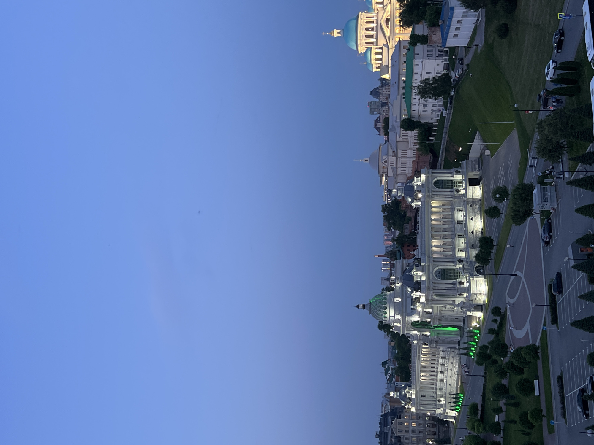 This is an eye-level, outdoor, rotated shot of the city of Kazan, Russia. Buildings are lit up at dusk, lining the right side of the frame. A street runs parallel to the buildings with greenery separating them. A parking lot is to the right of the street. A car is parked and another car is driving on the street. The sky is a gradient of dark and light blue on the left side of the image. The agricultural palace of Kazan and the Kremlin can be seen in the image.