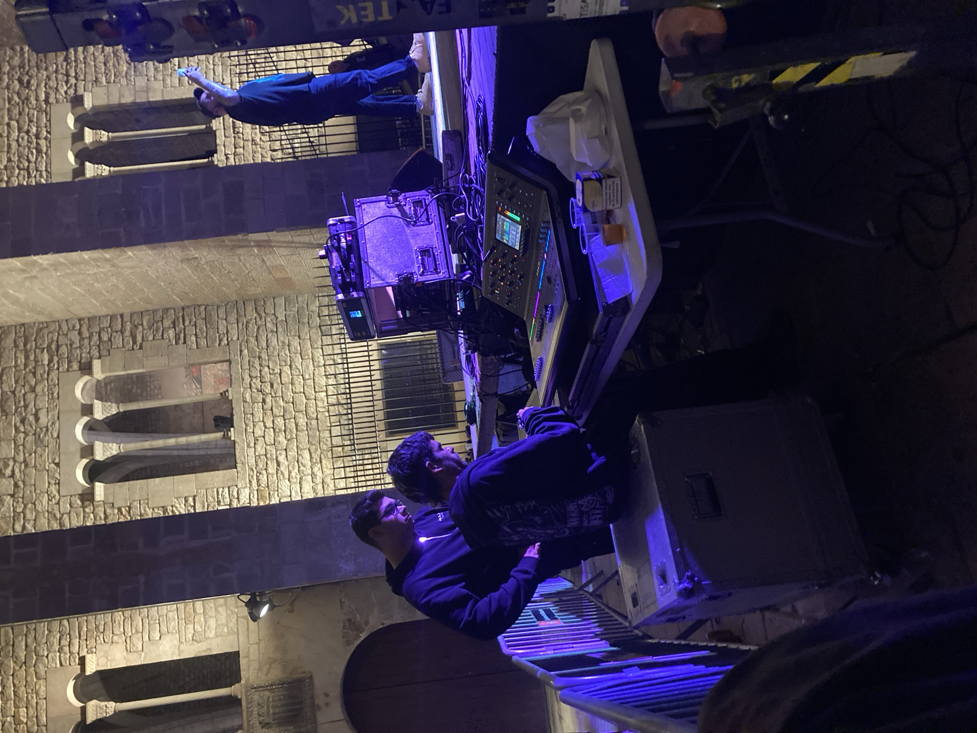 Two people stand behind a sound mixing console set on a table. The table also holds a case and some bottles. The console is lit with blue and purple light, as is the grating underneath the people. Above them is a man in blue clothes pointing to something out of frame. A stone building facade is visible in the background with arched windows. A sign says "FA TEK" at the top of the frame. The location may be in Barcelona, Spain, based on the architectural style.