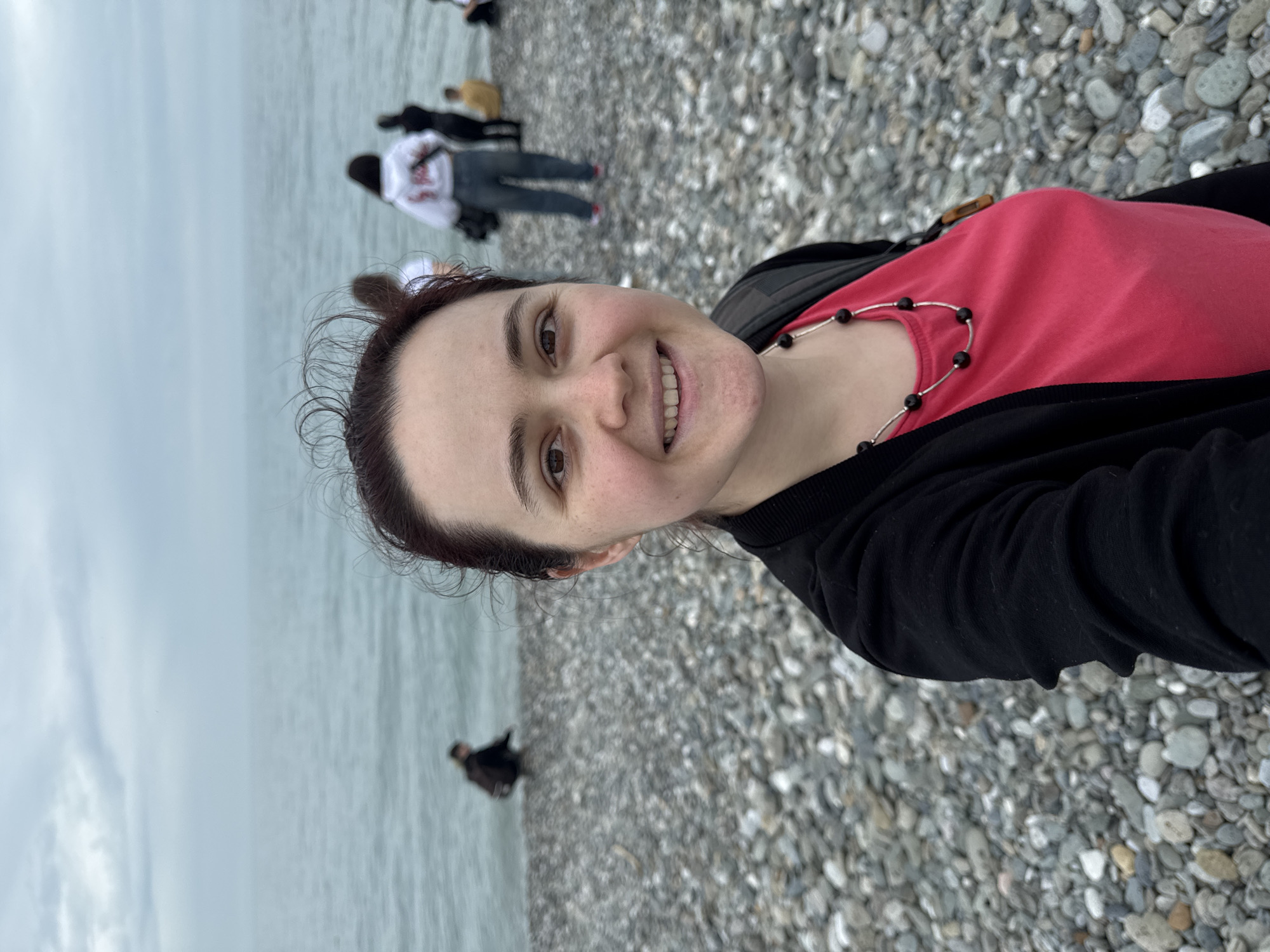A woman is taking a selfie on a pebble beach. The woman is wearing a red t-shirt, a black sweater, and a black beaded necklace. She is smiling and looking at the camera. In the background, there are several people walking along the beach and standing in the water. The sea is calm and gray. The sky is overcast. The location is likely a beach in Sochi, Russia, given the image's origin.
