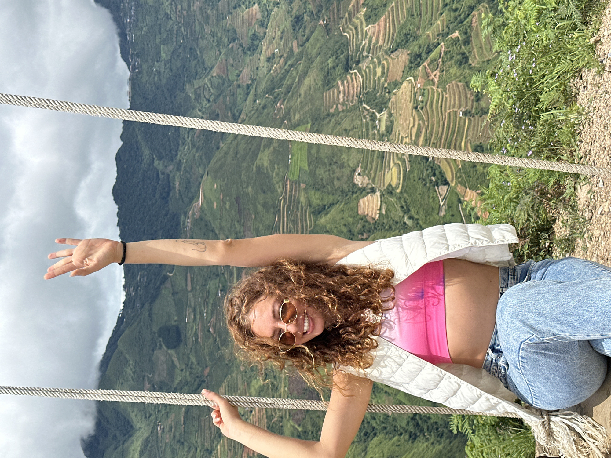 A young woman with curly brown hair, wearing sunglasses, a pink crop top, a white vest, and light blue jeans, is sitting on a swing overlooking a mountain landscape with terraced fields. Her arms are raised, and she is smiling. The scene is set in Ta Xua, Vietnam, known for its stunning natural beauty and rice terraces. The weather appears to be overcast with clouds obscuring the mountain peaks.
