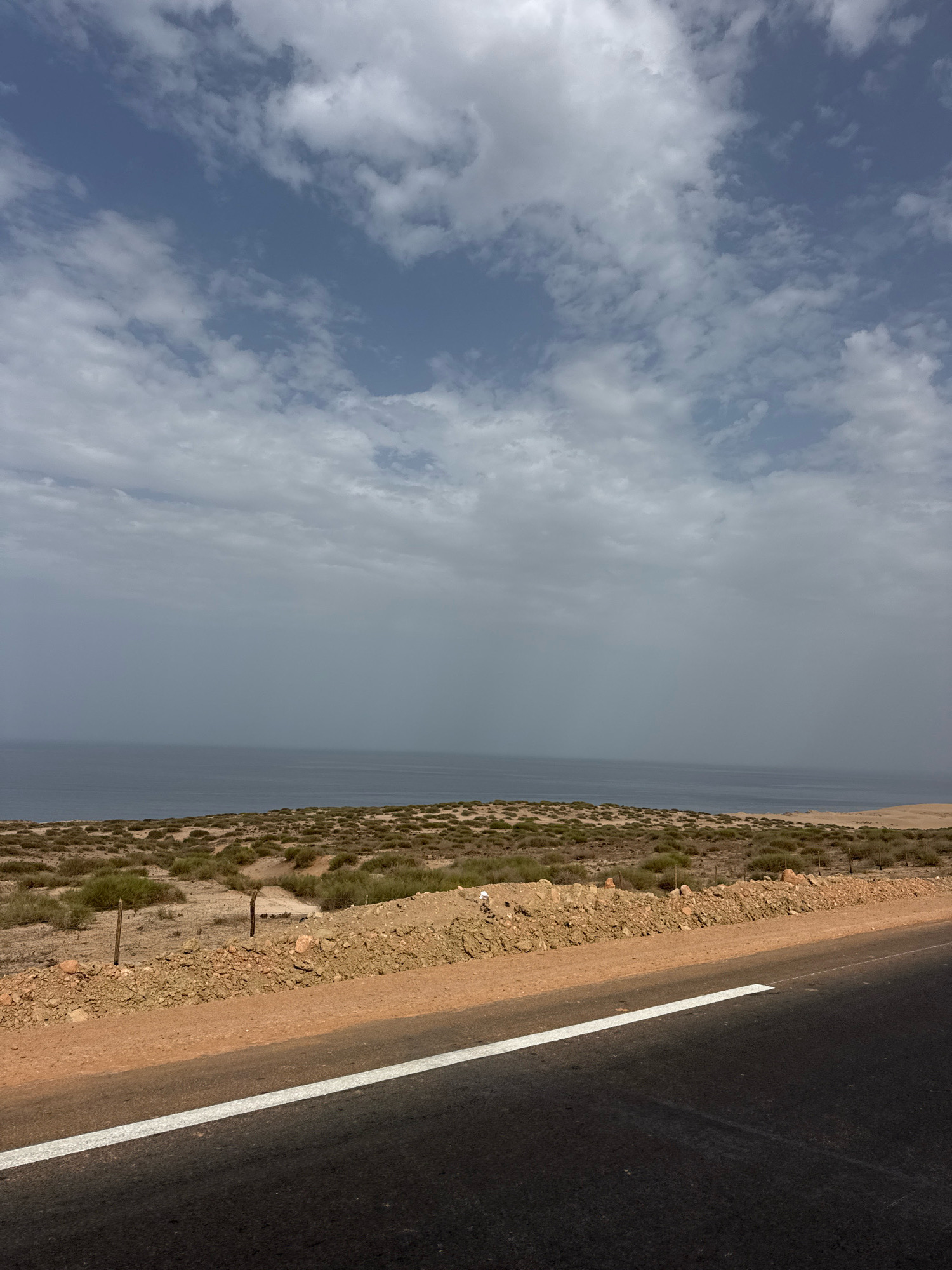 The image depicts a coastal landscape with a road in the foreground. The road is asphalt with a white line marking its edge. Beyond the road, there is a strip of dry, sandy ground with sparse, low-lying vegetation, characteristic of the arid coastal regions of Tamri, Morocco. This area appears somewhat barren with a few wooden posts visible, suggesting a boundary or fence. Further in the background, a vast expanse of the Atlantic Ocean stretches out, its surface appearing calm and slightly hazy under a cloudy sky. The clouds are varied, with some patches of blue sky visible, indicating a partly cloudy day. The overall impression is one of a remote, coastal road with a rugged, natural environment.