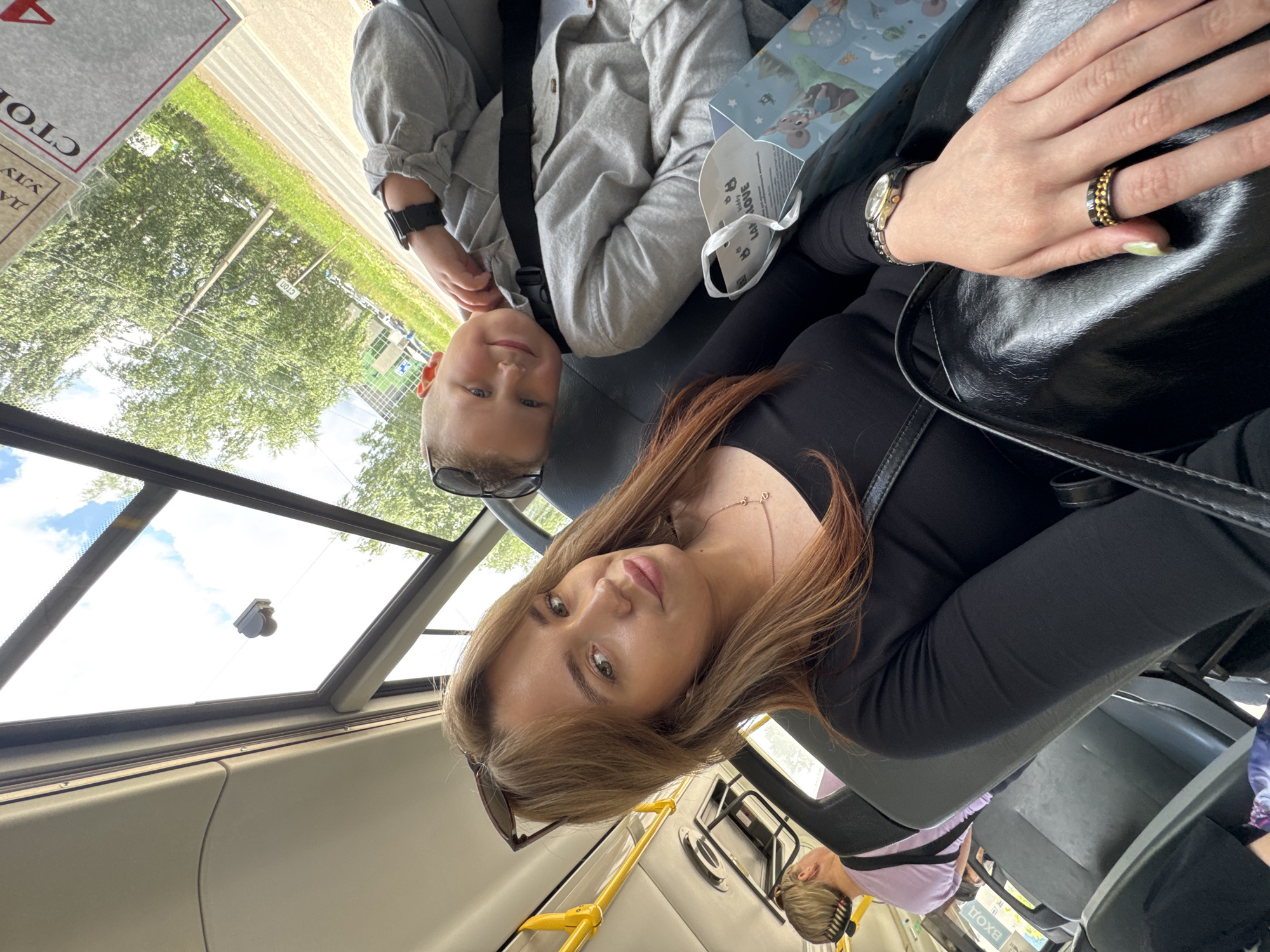 The image depicts a woman and a young boy taking a selfie on a bus, likely in Chaikovsky, Russia. The woman is in the foreground, facing the camera with her head slightly tilted. She has long, brown hair with reddish highlights and is wearing a black long-sleeved shirt and sunglasses perched on her head. She is also wearing jewelry on her right hand, including a ring with gold and black elements and a wristwatch. To her right and slightly behind her, a young boy is visible. He is wearing a light grey hooded jacket and sunglasses. He is looking towards the camera with a smile, and his arm is resting on his chest. In the background, other passengers are seated, and the interior of the bus, including yellow grab poles and dark grey seats, is visible. Through the windows, trees and a cloudy sky can be seen, suggesting an outdoor environment. A sign with Cyrillic text, possibly indicating a bus stop or route information, is also visible on the bus. The overall scene suggests a casual journey on public transportation.