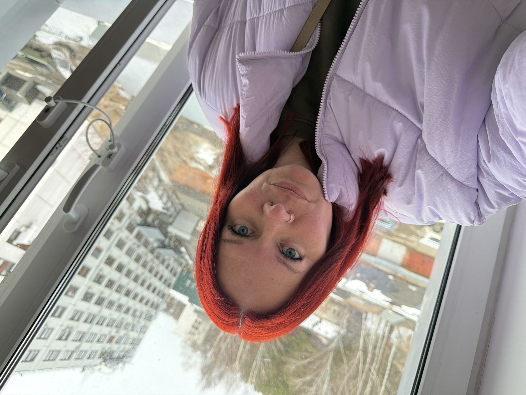 The image displays a close-up portrait of a female individual captured indoors, looking out through a window onto an urban environment in Chaikovsky, Russia. The subject, a woman in her 20s to 30s, possesses bright red hair with a visible part and blue eyes. A silver-colored hoop nose ring is present on her left nostril. She is wearing a light purple quilted puffer jacket over an olive-green top, with a light-colored strap visible across her right shoulder. Her expression is neutral as she directly faces the camera.
The immediate foreground includes a white multi-section window frame, featuring a white handle on the right side and a white safety cable connecting two points of the frame. Beyond the window, the background reveals an overcast urban landscape, indicative of late winter or early spring conditions. Bare deciduous trees are visible in the mid-distance on the left, and patches of dirty snow are present on the ground. A prominent light-colored, multi-story building with multiple windows dominates the right side of the external view. To the left, several lower-rise structures, resembling industrial buildings or garages, are visible, alongside indistinct vehicles in the distance, including what appears to be a white van or car. The perspective suggests an elevated vantage point.