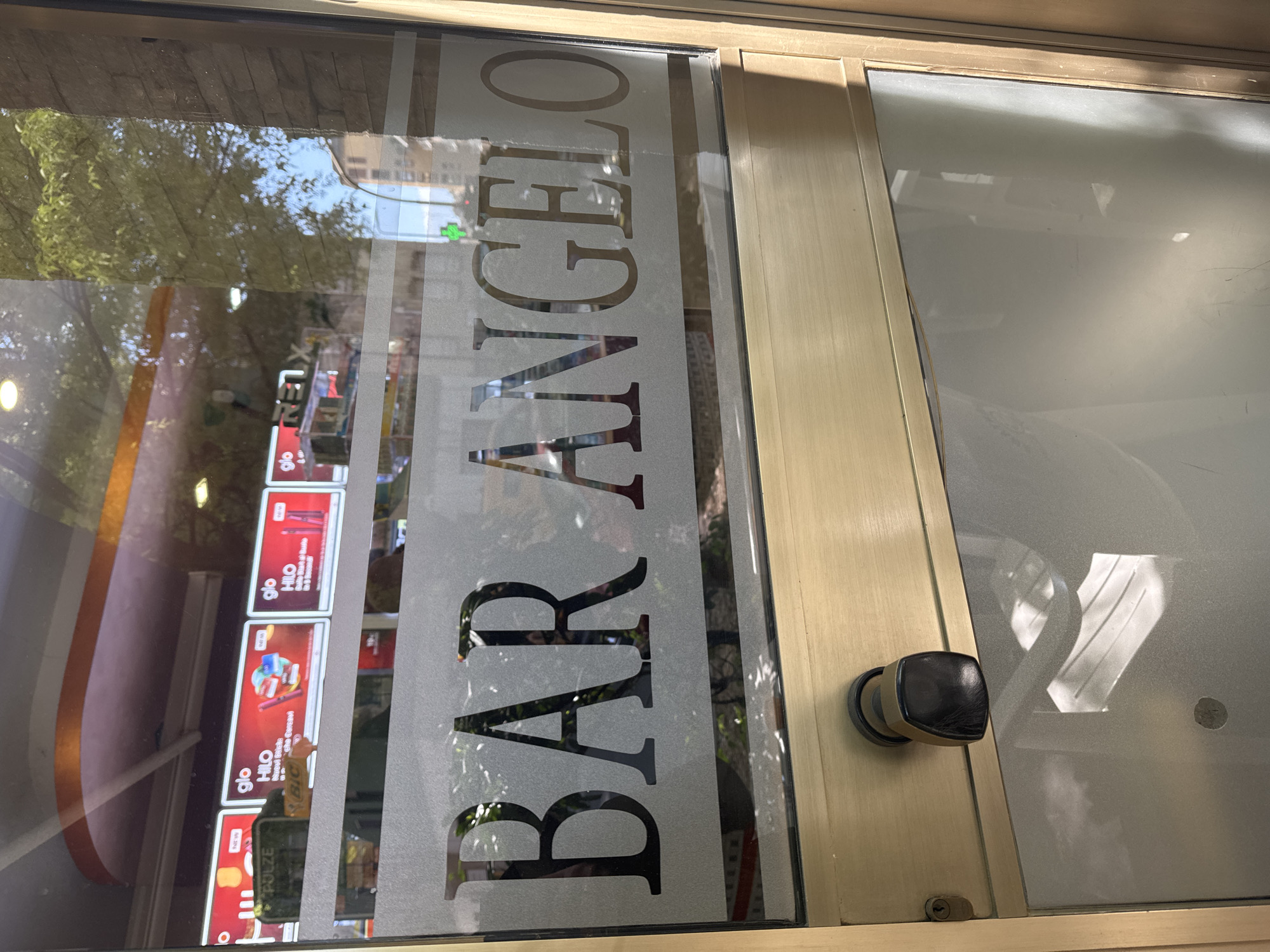 A storefront in Milan, Italy displays the words "BAR ANGELO" vertically on its glass entrance door. The door also features a black, rounded handle. The glass door is set within a gold-colored frame. Through the glass, promotional posters are visible inside, featuring the word "glo" and depictions of product advertisements. A green pharmacy cross and buildings are reflected in the glass, suggesting an urban environment.