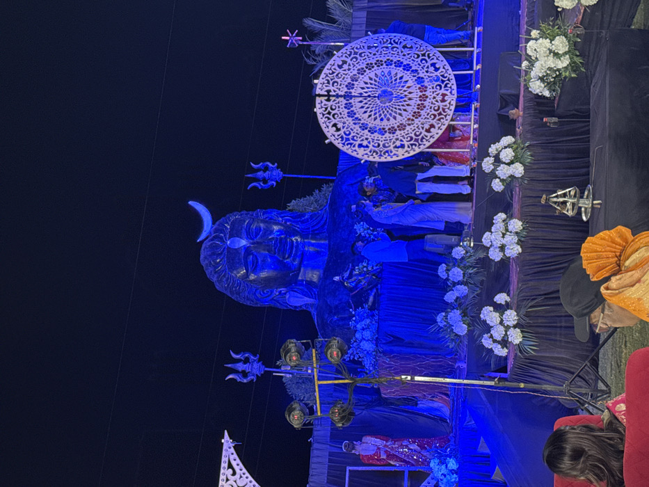 An outdoor night scene features a large, blue-lit statue of a deity with a crescent moon above its head. Floral decorations and patterned circular ornaments adorn the space. People wearing traditional attire stand on a platform behind the statue. Below, individuals are seated, with one wearing an orange turban. The overall scene suggests a cultural celebration or religious event.