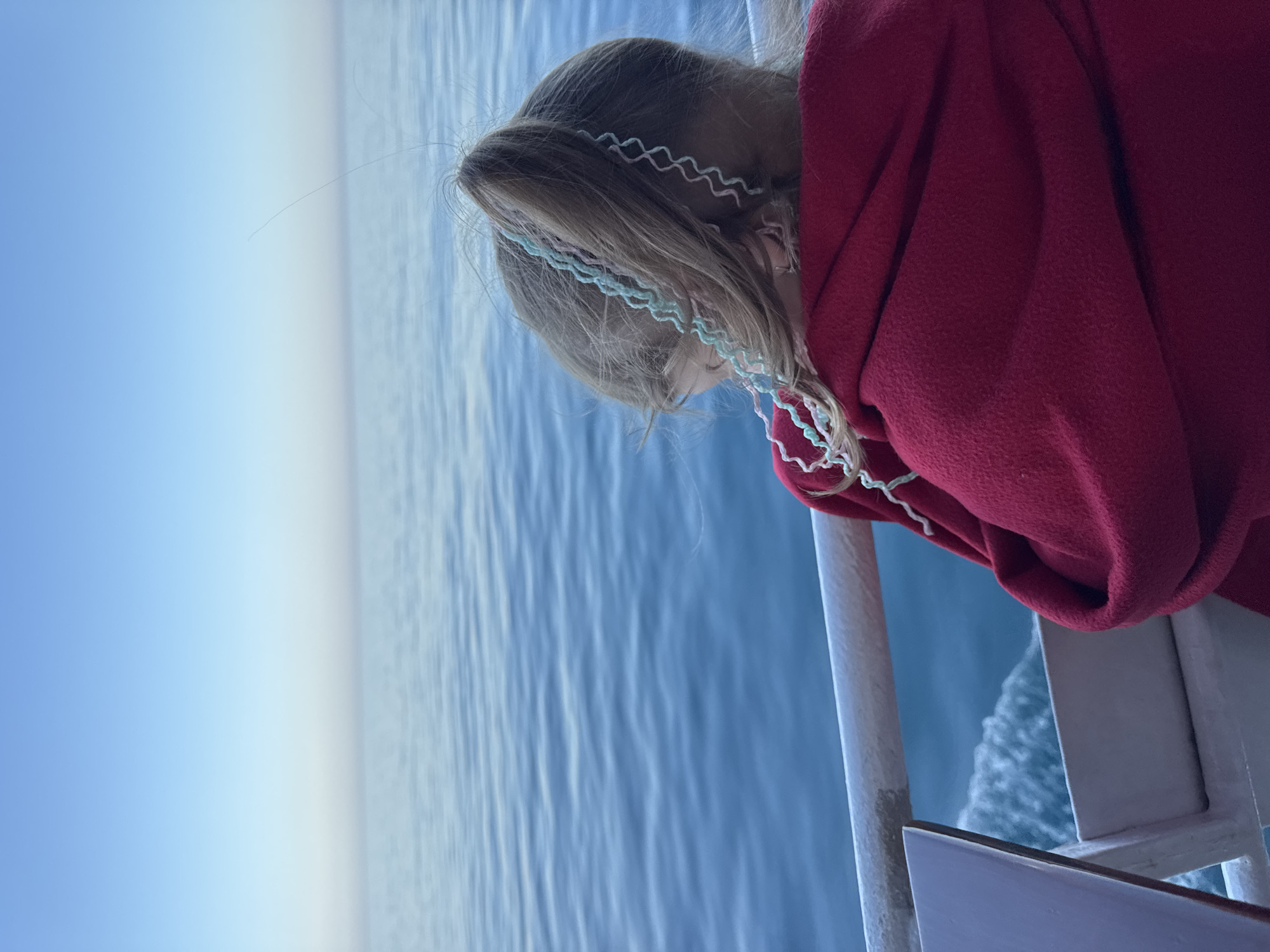 A young girl with blonde hair wearing a red sweater leans over the railing of a boat. The boat is at sea, city unknown.