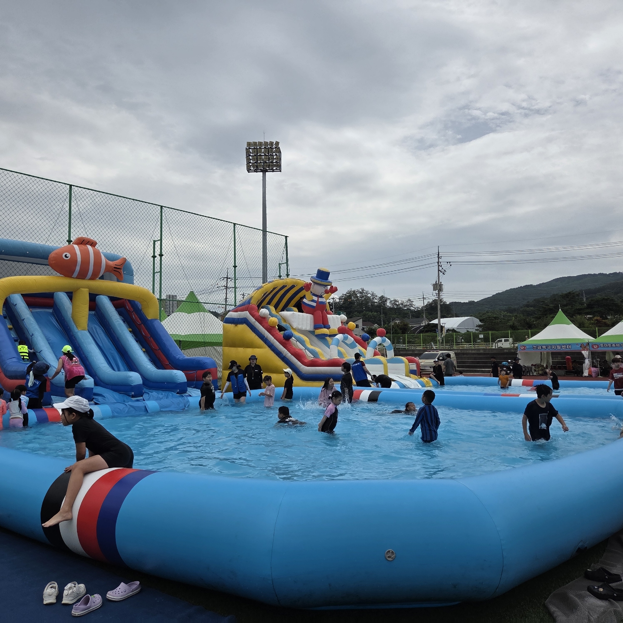 In Dunnae-myeon, South Korea, a family-friendly inflatable water park is depicted on an overcast day. A large, blue inflatable pool is the central focus, filled with clear water. Several children are actively playing in the water, some on the inflatable slides and others wading or swimming. A prominent inflatable water slide complex features multiple blue slides, a large orange fish decoration at the top, and a clown-like figure. The inflatable structure is multicolored with yellow, blue, red, and white sections. A chain-link fence with green posts encloses the area, with a tall stadium light visible in the background. In the distance, there are green tents, a street with parked vehicles, houses, and tree-covered hills under a cloudy sky. On the edge of the pool, a young girl in a black shirt and shorts sits on the inflatable barrier, her bare feet resting on it. Several pairs of shoes, including white sneakers and purple Crocs, are placed on the blue matting beside the pool.