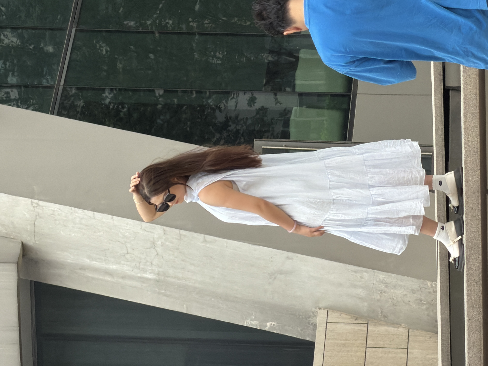 In Yuzhong District, China, a young woman stands on a concrete staircase, her back to the camera, facing a building with a textured concrete facade and dark green glass windows. She wears a light blue tiered dress, white socks, and black and white loafers with pearl accents. Her right hand is raised to her head, and she sports sunglasses. To her right, the back of a man in a blue t-shirt is visible, also facing away from the camera. The environment suggests an urban setting with modern architecture.