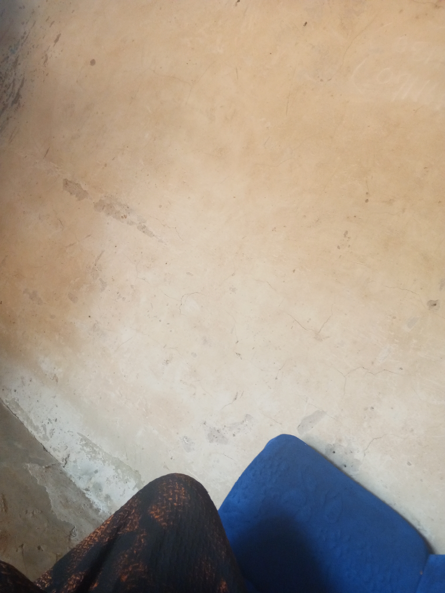 The image shows a close-up view of a concrete floor with a pattern of stains and cracks. In the lower right corner, a section of a bright blue upholstered surface, possibly a chair or sofa, is visible. In the lower left corner, a portion of dark fabric with an orange and black pattern is also visible, suggesting the presence of a person or an item of clothing. The lighting is diffused, creating subtle shadows and highlighting the texture of the concrete. There is no specific indication of the location within the city of Nyabe, Nigeria, other than the general appearance of a rustic or utilitarian indoor setting.