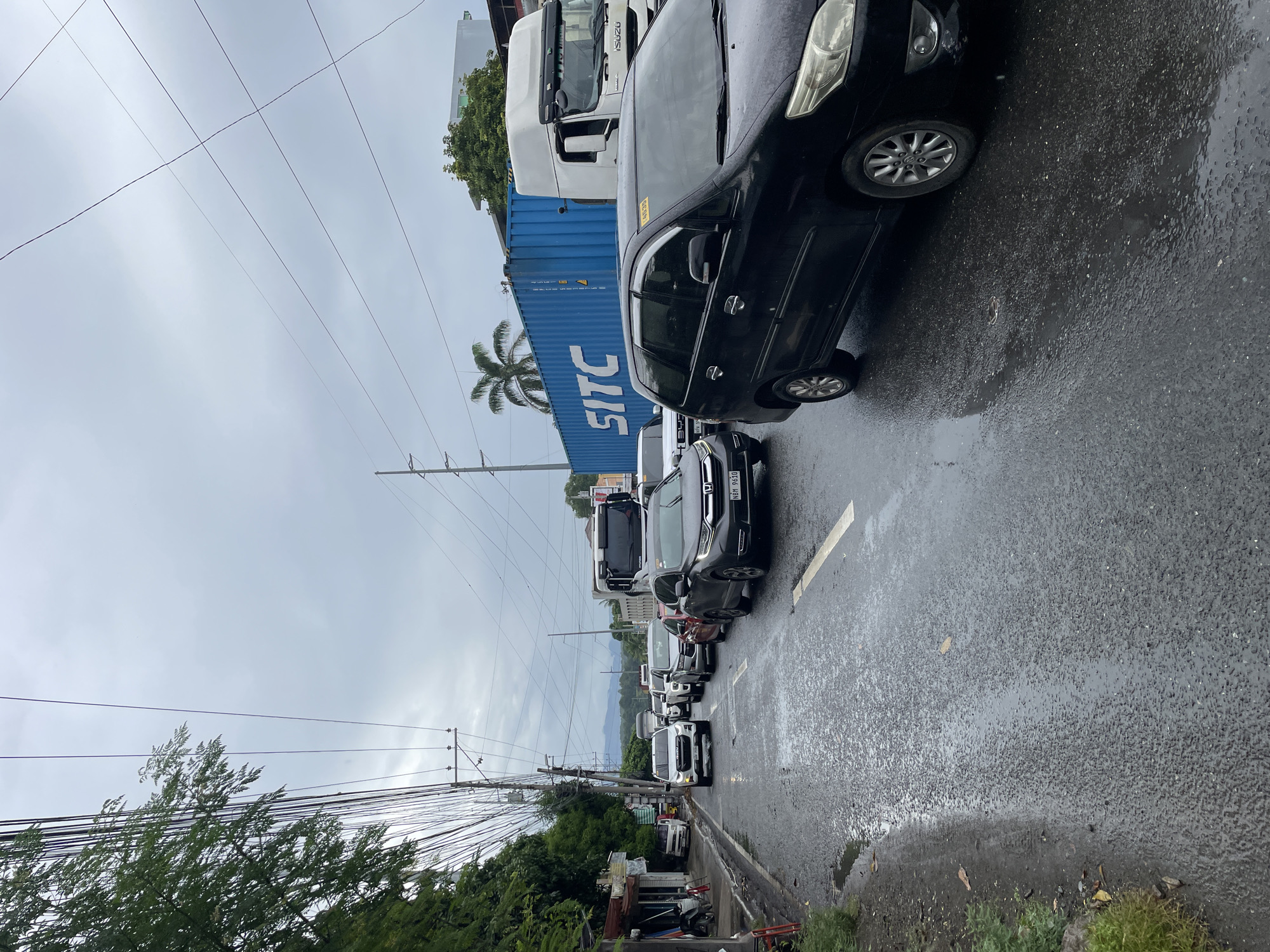 This is a vertical, eye-level shot of a traffic jam on a wet asphalt road in Davao City, Philippines. The sky is overcast and grey. In the foreground, a black SUV is stopped in traffic, with its right front wheel visible. Further up the road, a white SUV with an "ISUZU" logo is also stopped. Behind it, several other vehicles, including cars and a truck, are visible, all appearing to be part of the traffic congestion. To the left of the road, there are some small shops or stalls and a row of trees. A large blue shipping container with "SITC" written on it is visible in the background, along with a white truck. The road is reflecting the grey sky and the vehicles due to the wet surface, with several puddles visible. The scene depicts a typical urban traffic situation in Davao City.
