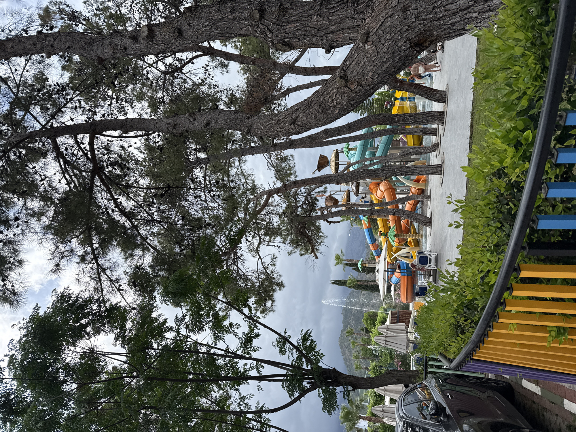 The photograph, taken in Kemer, Turkey, shows a waterside recreation area under cloudy skies. In the background, a mountain range is partially obscured by clouds. A water fountain shoots upward in the middle ground. Water slides of varying colors (blue, yellow, orange) are visible. There are trees around the perimeter of the area. A dark-colored car and a colorful fence (blue, yellow, purple) are in the foreground.
