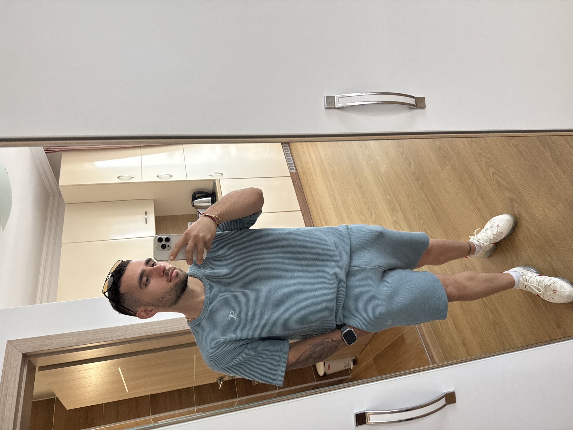 A young man is taking a mirror selfie in a domestic setting. He is wearing a light blue short-sleeved t-shirt and matching shorts. The t-shirt has a small white logo on the left chest. He has short dark hair, a beard, and is wearing sunglasses on his head. He has a watch on his left wrist and a bracelet on his right wrist. His hands are in his pockets. He is standing on a wooden floor in front of a full-length mirror. To the left of the mirror, there is a glimpse of a bathroom with a trash can. Behind him, in the reflection, a kitchen area is visible with light-colored cabinets and a kettle on the counter. The overall environment suggests an apartment or house interior. The scene is well-lit. The city of Nesebar, Bulgaria is not depicted in this image.