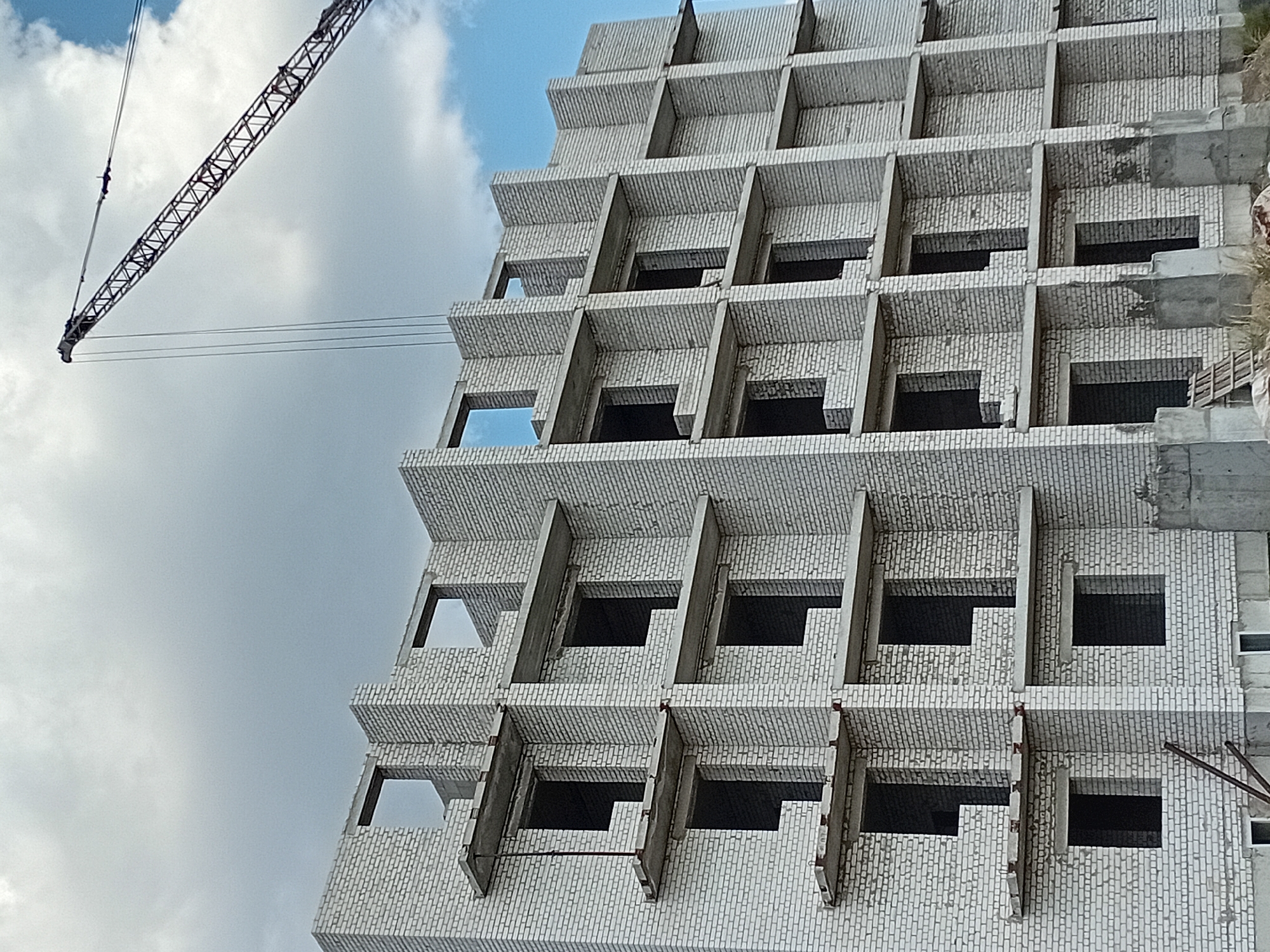 A low-angle, full shot captures a multi-story building under construction in an outdoor setting. The building is constructed of white bricks, with numerous open window frames and concrete balconies. A large construction crane with a segmented boom is visible in the upper right quadrant of the image, extending over the building and into the cloudy sky. The lower portion of the building shows concrete foundations and some scaffolding. The sky is filled with a mix of gray and white clouds, with a hint of blue sky peeking through. The scene is devoid of people.