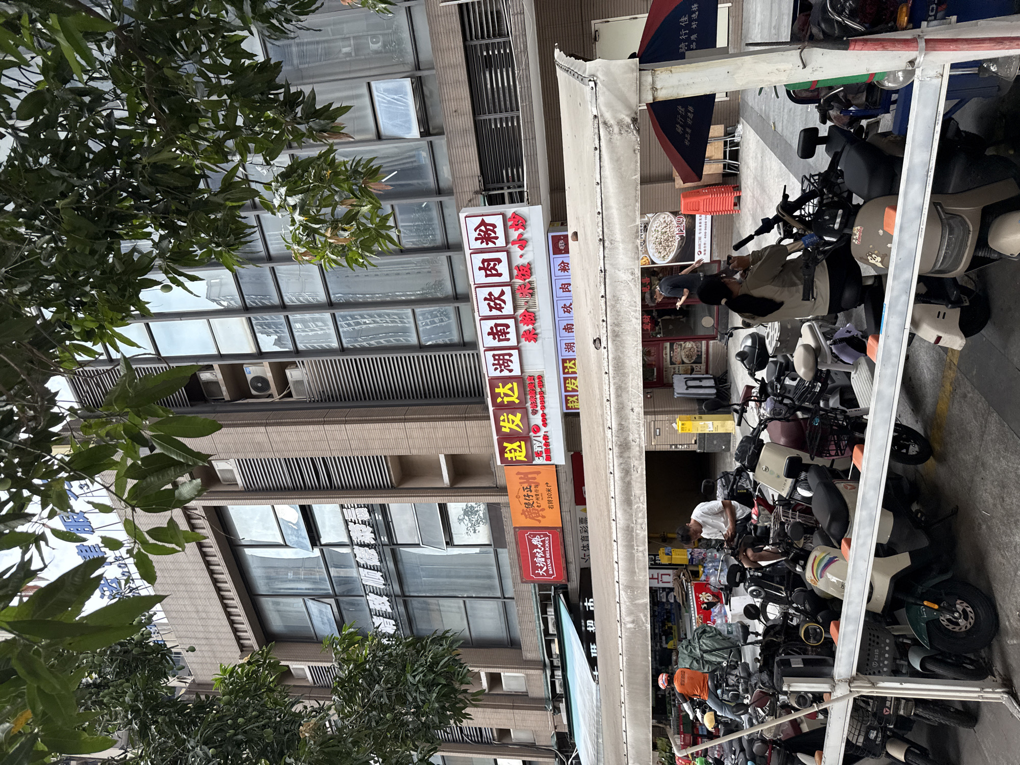 In Haizhu District, China, a street scene unfolds beneath a canopy, revealing a collection of parked scooters and motorcycles. A person in an orange shirt rides a scooter away from the viewer, while another individual, dressed in a white t-shirt, sits on a motorcycle engrossed in their phone. A woman with her hair in a ponytail is seated on a scooter, holding a phone to her face as if taking a photo. The background displays a multi-story building with various shops on the ground floor, characterized by red and white signage. One sign prominently features Chinese characters and the text "BATANG DELICIOUS." The overall environment suggests a bustling urban commercial area.