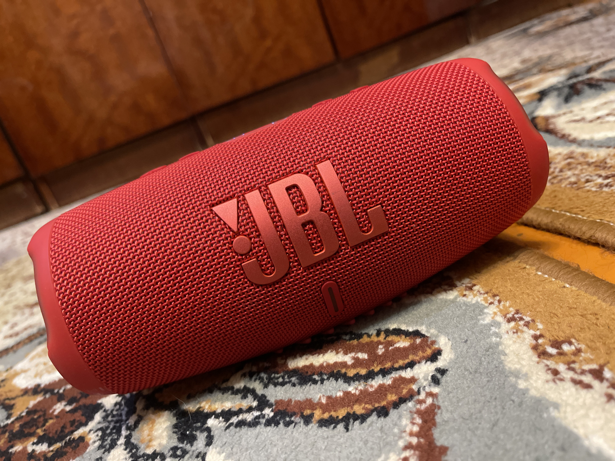A close-up, angled shot shows a red JBL portable speaker resting on a carpet with a brown and tan patterned design. The speaker is cylindrical with a textured fabric exterior. The JBL logo is prominently displayed in a raised, metallic red on the side of the speaker. The background is blurred and appears to be a wooden surface. The overall scene suggests an indoor setting, possibly a home. There is no indication of the city of Bogdanovich, Russia, in the image.