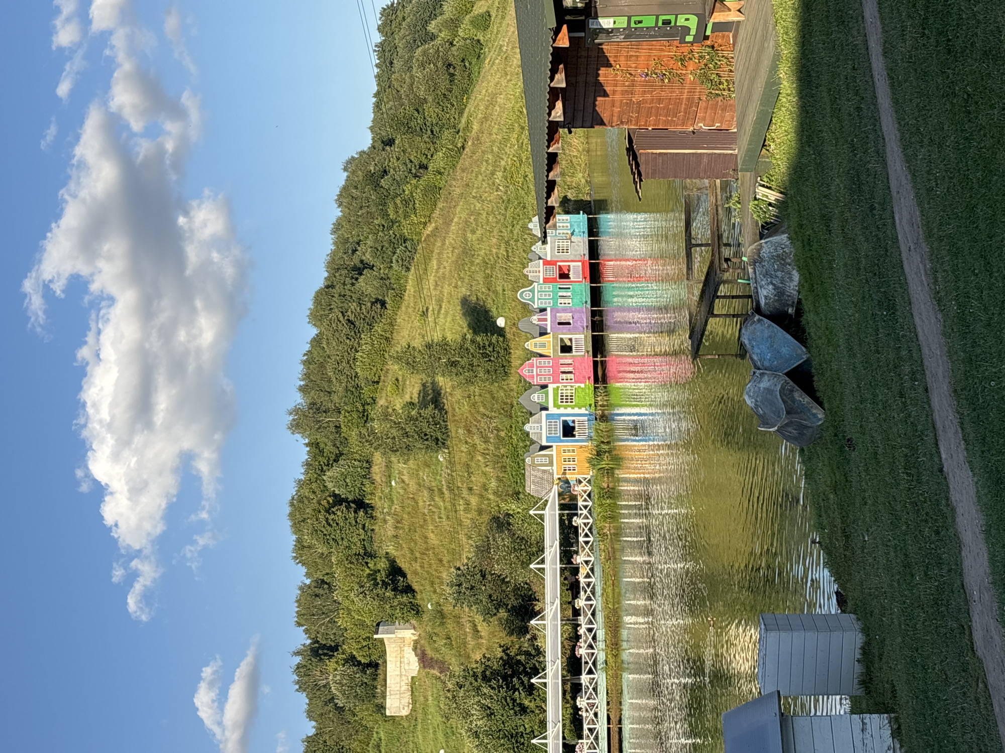 The image captures a scenic view of a lake in Romashkovo, Russia, on a clear day with scattered clouds. In the foreground, a grassy bank is visible with two small, white structures and several overturned boats. A wooden deck and a small wooden building are situated to the right of the frame. The lake itself is calm, reflecting the vibrant colors of a row of small, colorful houses that appear to be floating on the water, resembling a miniature Dutch village. These houses are painted in bright hues of pink, purple, green, blue, and yellow, each with white trim and small windows. Behind the floating houses, a white awning stretches over a walkway or patio area, with tables and chairs set up, suggesting a dining or recreational space. The background is dominated by a lush, green hillside covered in trees and vegetation. A concrete structure is partially visible on the hillside. The overall impression is one of a tranquil and picturesque lakeside resort or park.