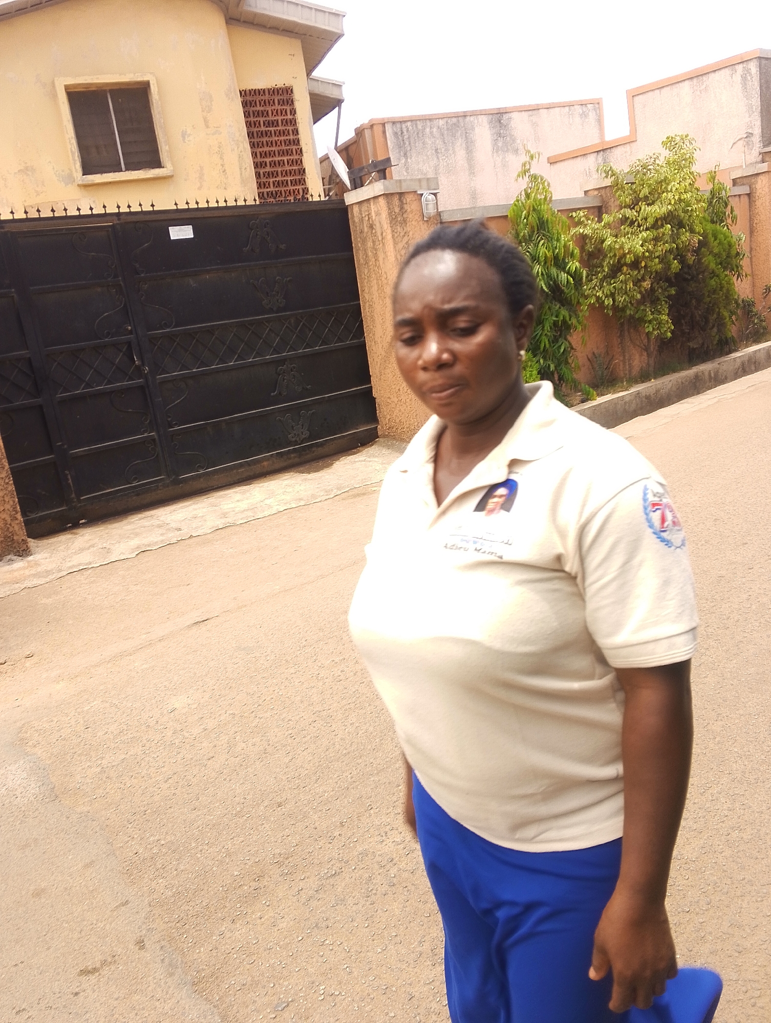 A woman stands on a paved street in front of a metal gate and building. The building is light yellow and has a small barred window. The metal gate is black. The woman has dark skin and dark hair pulled back. She wears a beige polo shirt with writing and a photograph on the front and a blue emblem on the sleeve. She also wears blue pants and carries a blue object in her hand. Behind her, a light brown wall surrounds greenery and a tree. The location is Jos, Nigeria.