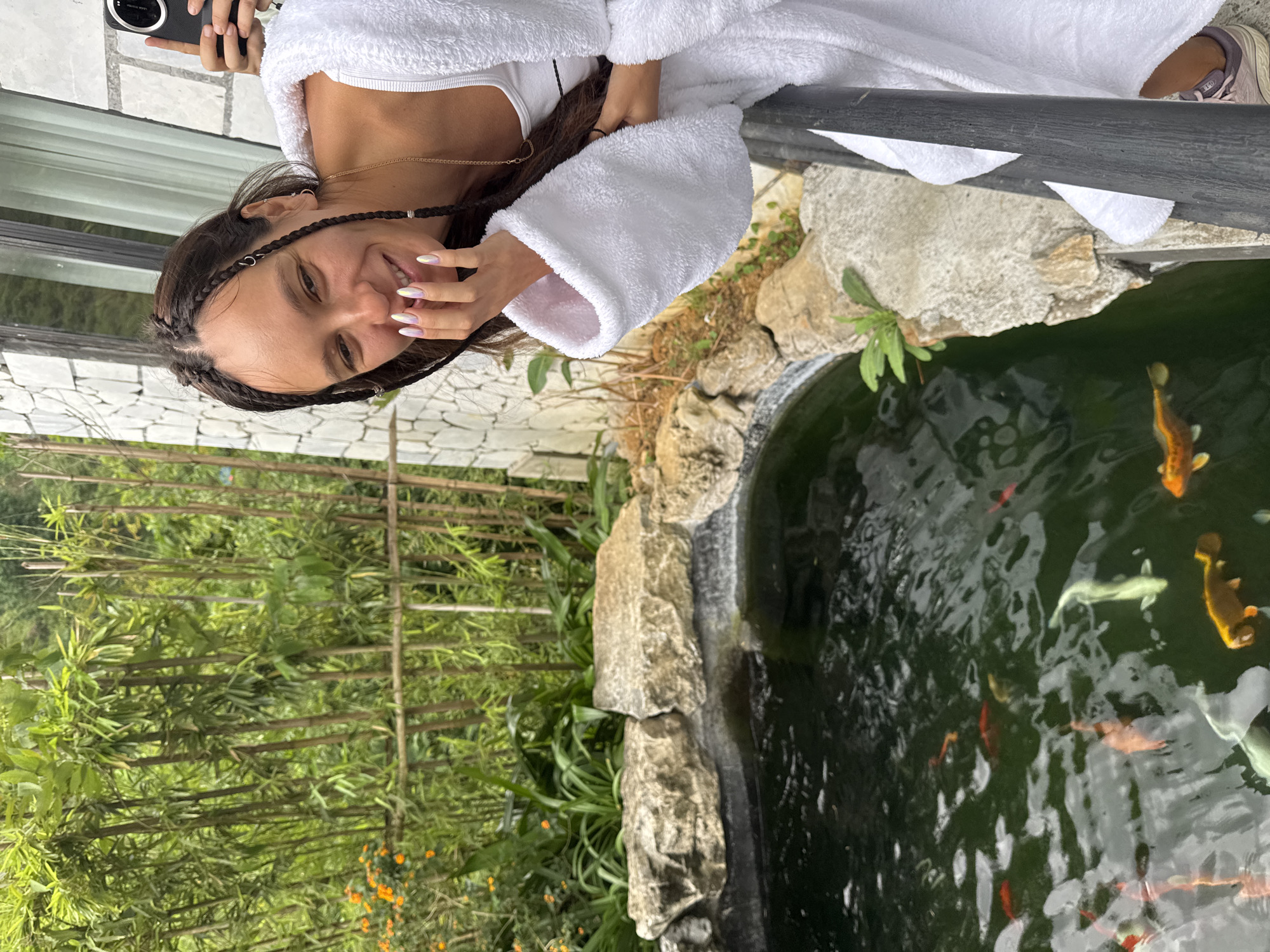 A young woman, likely in her early 20s, with dark hair braided on the sides, stands next to a pond filled with koi fish. She is wearing a white bathrobe and white athletic top underneath and is holding a cell phone. Her nails are manicured with a holographic design. The background shows a garden setting with bamboo and other lush greenery and bamboo fencing. The location appears to be Cat Cat Village, Vietnam, as the pond's surroundings, vegetation, and the stone and white-washed building behind her are consistent with the area's landscape and architecture. The pond is bordered by large rocks, suggesting a naturalistic or landscaped setting.