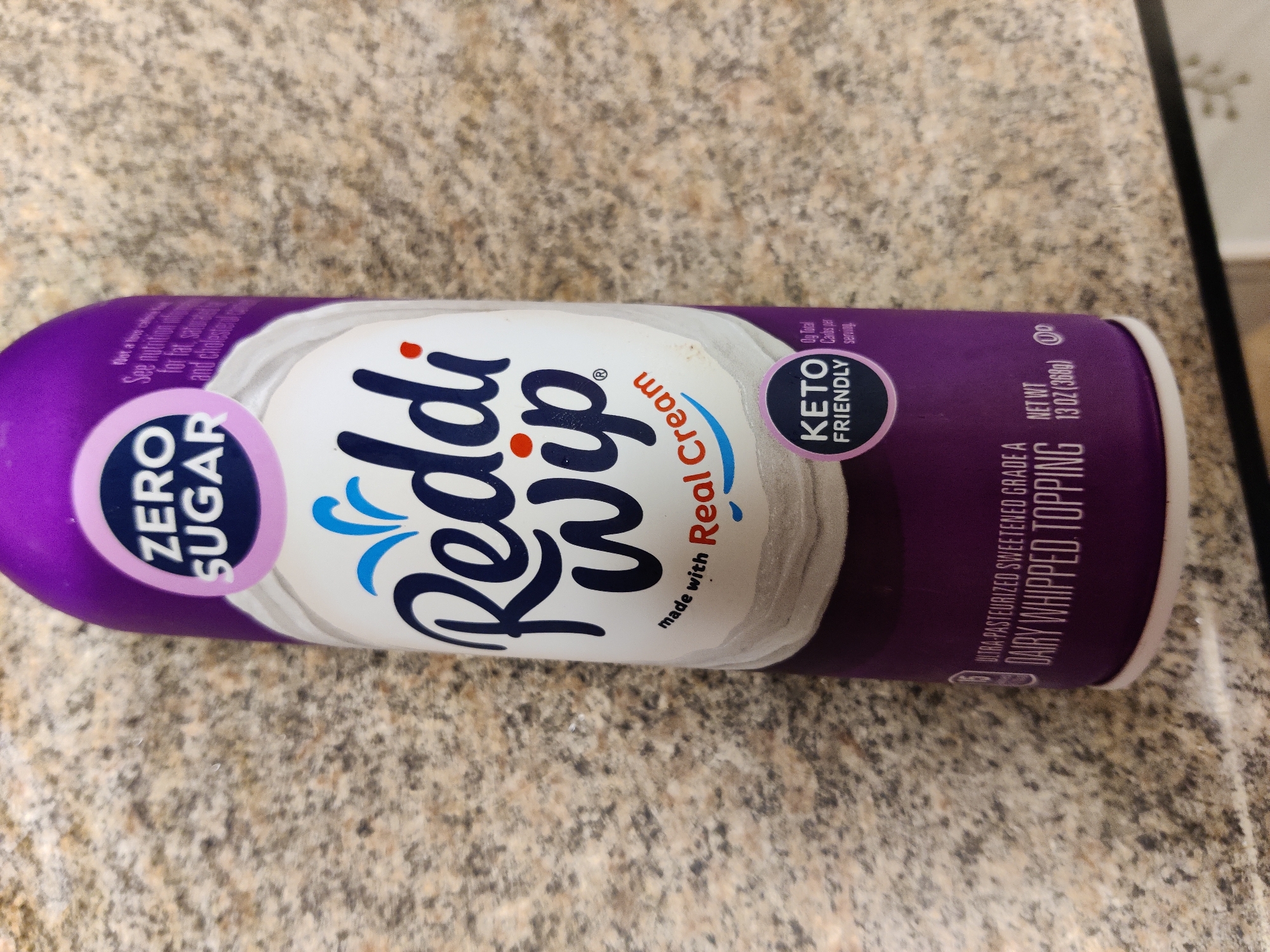 A close-up, slightly angled, horizontal shot features a can of Reddi-wip dairy whipped topping. The can is primarily purple and white with dark blue lettering. On the left side of the can, a circular purple label contains white text reading "ZERO SUGAR". The brand name "Reddi" is written in large, dark blue script across the center of the can, followed by "wip" in similar lettering. Above "wip", a blue wave design is present. To the right of "wip", a label reads "made with Real Cream". A smaller, round, purple and pink label on the right side of the can states "KETO FRIENDLY". Further to the right, smaller white text on the purple background reads "ULTRA-PASTEURIZED SWEETENED GRADE A DAIRY WHIPPED TOPPING" and "NET WT 13 OZ (368g)". The can rests on a speckled beige and gray countertop.