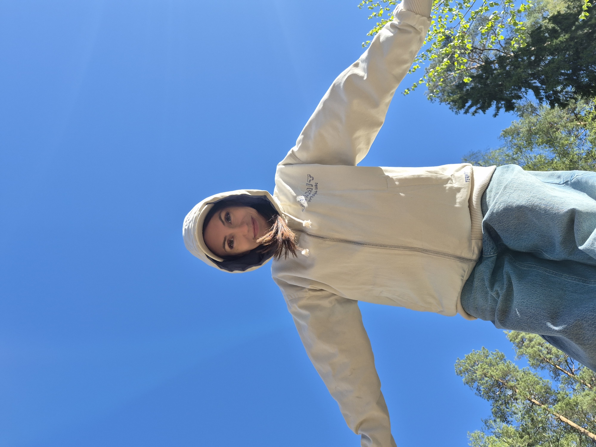 A low-angle shot shows a woman standing outside in Chaikovsky, Russia, against a bright blue sky. She wears a light-colored hooded jacket and blue jeans. Her arms are outstretched, and she looks down at the camera with a slight smile. Tree branches with green leaves are visible in the upper right and lower right corners of the frame.