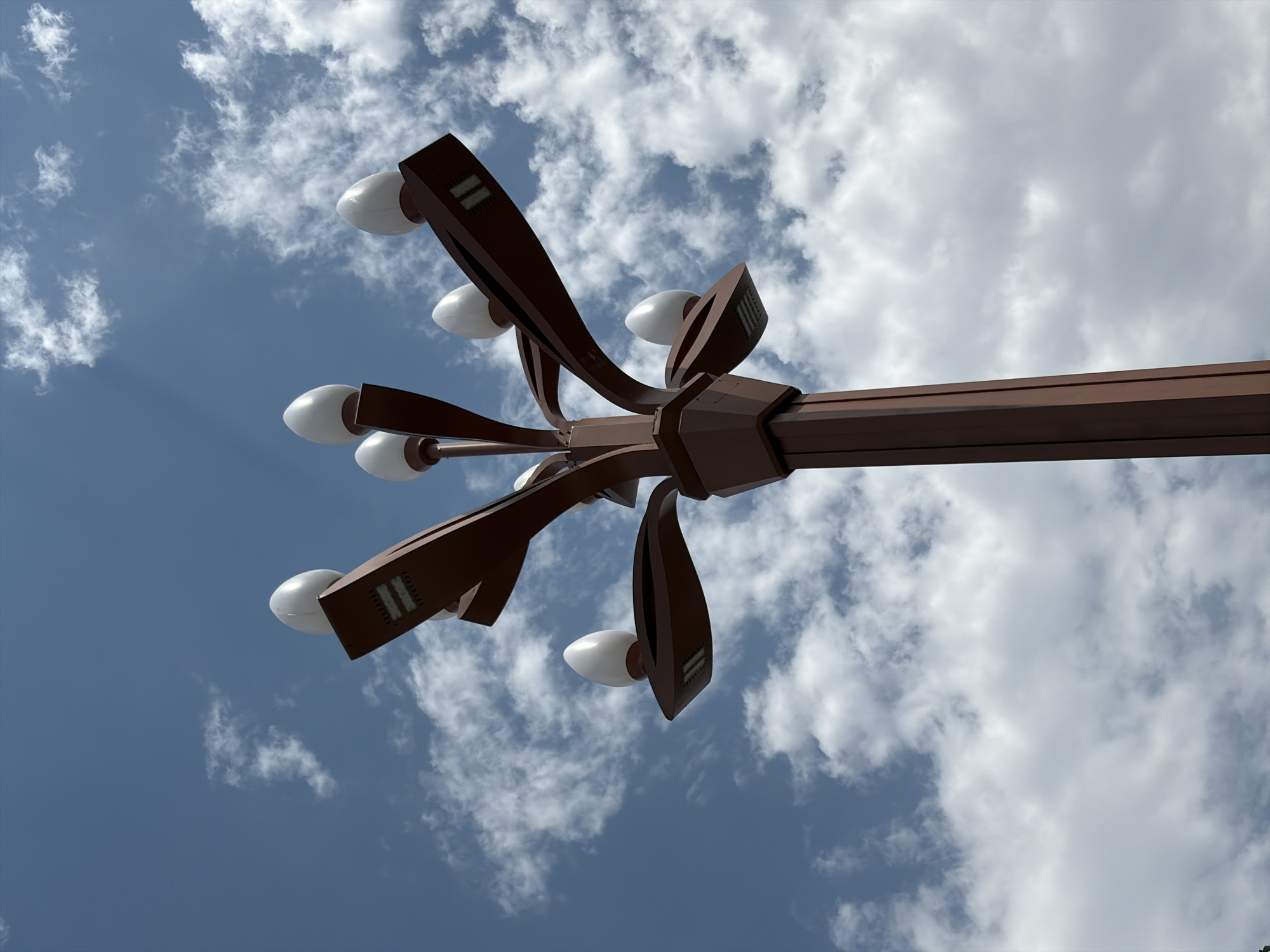 A close-up, low-angle shot features a brown, multi-tiered lamppost with egg-shaped white light fixtures against a bright blue sky with scattered white clouds. The lamppost has a central, thick vertical post from which several arms extend outward, each holding a light. The light fixtures appear to be modern, with a sleek, elongated shape and visible LED strips beneath the opaque covers. The sky is partially obscured by the lamppost and its branches, with thin streaks of sunlight visible through the clouds in the upper left quadrant. The overall impression is one of a clear, bright day in an urban or park setting. The image does not contain any people or discernable actions. The specific location within Bishkek City, Kyrgyzstan, is not identifiable from the visual information present.