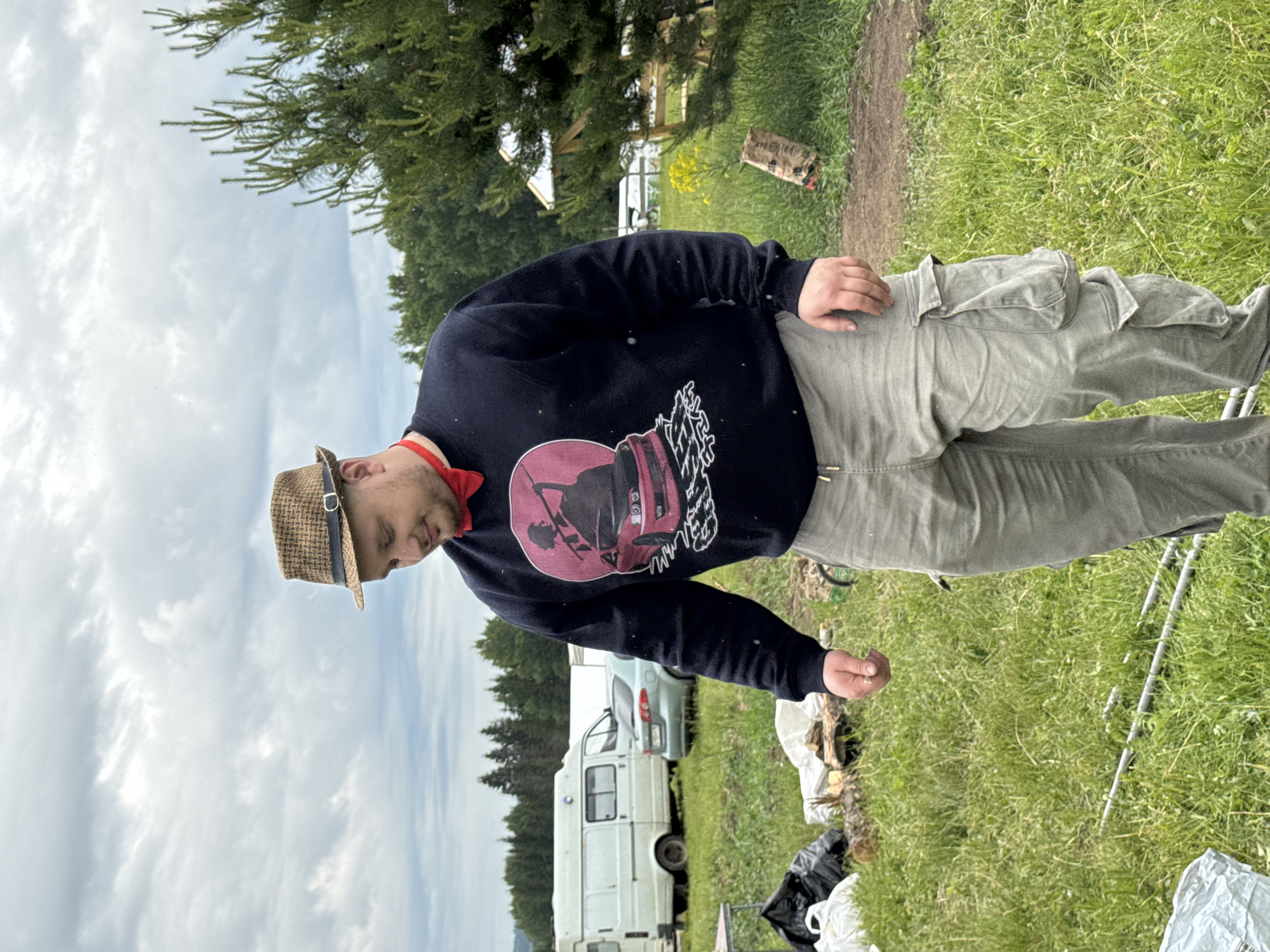 The image, taken in Bol'shaya Kivara, Russia, shows a man standing outdoors on an overcast day. He is wearing a straw fedora-style hat, a red bandana, a dark blue or black long-sleeved sweatshirt, and light-colored cargo pants. The sweatshirt features a prominent graphic of a stylized car, rendered in shades of red and pink with white outlines and abstract, illegible text or shapes above and below it.
The man is positioned slightly to the left of the frame, looking downwards and to his right. His left hand rests on his hip or pocket, while his right hand is held out slightly, possibly holding a small, indistinct object.
The setting is a rural or semi-rural outdoor area. In the background, a large green conifer tree is visible. A narrow dirt path runs alongside a grassy area to the man's right. Further back, a white camper van and a light-colored car are parked. The ground is covered in tall green grass, with scattered debris, including pieces of wood and black and white bags, and some metal poles lying on the ground. The weather appears to be overcast during daylight hours.
