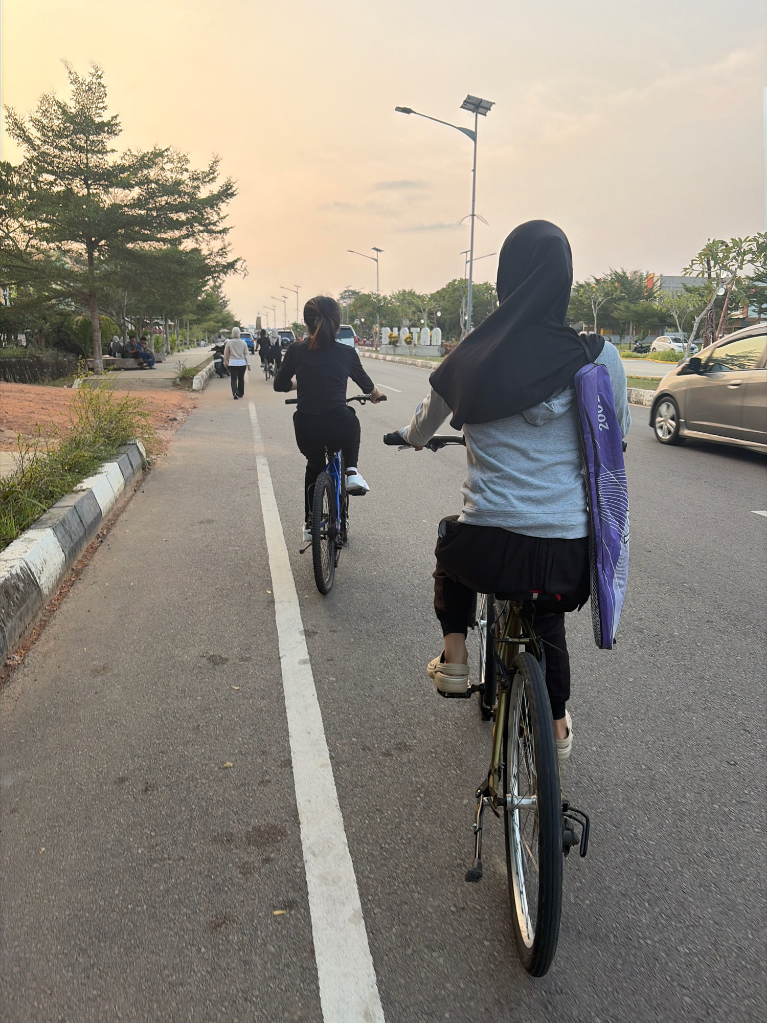In Tanjung Pinang, Indonesia, two individuals are cycling along an asphalt road during what appears to be the late afternoon or early evening, given the warm, soft lighting of the sky. The sky is a blend of pale orange and yellow, suggesting either sunrise or sunset, with some diffused clouds.
The main subject in the foreground is a person on a bicycle, viewed from behind. This cyclist is wearing a black hijab and a light gray hooded jacket over black pants. They are carrying a long, purple bag with white line designs strapped diagonally across their back, extending from the shoulder to the hip. The bag has "200" visible in white text at the top. They are wearing light-colored slip-on shoes.
Ahead of this cyclist is another person on a bicycle, also seen from behind. This individual is wearing a black long-sleeved shirt and black pants. They have dark hair tied back.
The road has a white line marking its center and a curb with black and white painted sections on the left. To the left of the road, there is a sidewalk or path lined with trees and some vegetation. A few people are seated on a bench or low wall under the trees. In the background, there are more trees, streetlights, and some buildings. A silver car is visible on the right side of the road, moving in the same direction as the cyclists. The overall scene conveys a sense of leisurely activity in an urban or suburban environment.