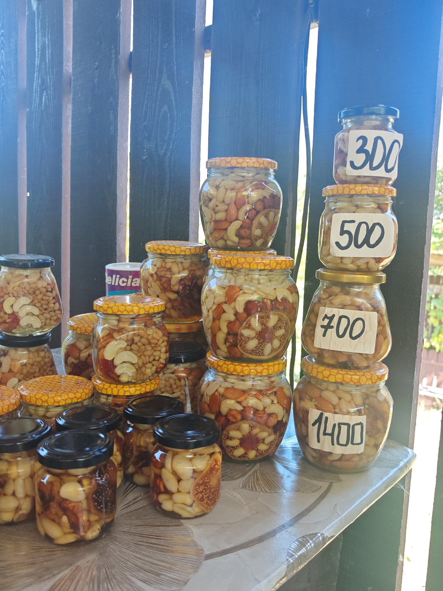 A close-up shot captures a stall displaying jars of nuts and dried figs, presumably for sale. The jars are of various sizes and are filled with a mixture of nuts, including almonds, cashews, and possibly walnuts, along with sliced dried figs. Some jars have yellow lids with a honeycomb pattern. Several jars have handwritten price tags: "300", "500", "700", and "1400". In the background, a dark wooden fence creates a backdrop, and out-of-focus greenery suggests an outdoor setting. The lighting indicates it's daytime, with sunlight casting shadows and highlights. A small purple and white can with the word "delicia" is visible behind some of the jars. The overall impression is that of a small, informal market stall, likely in a rural or semi-rural area, possibly in or near the village of Varvarovka, Russia, given the specific context.