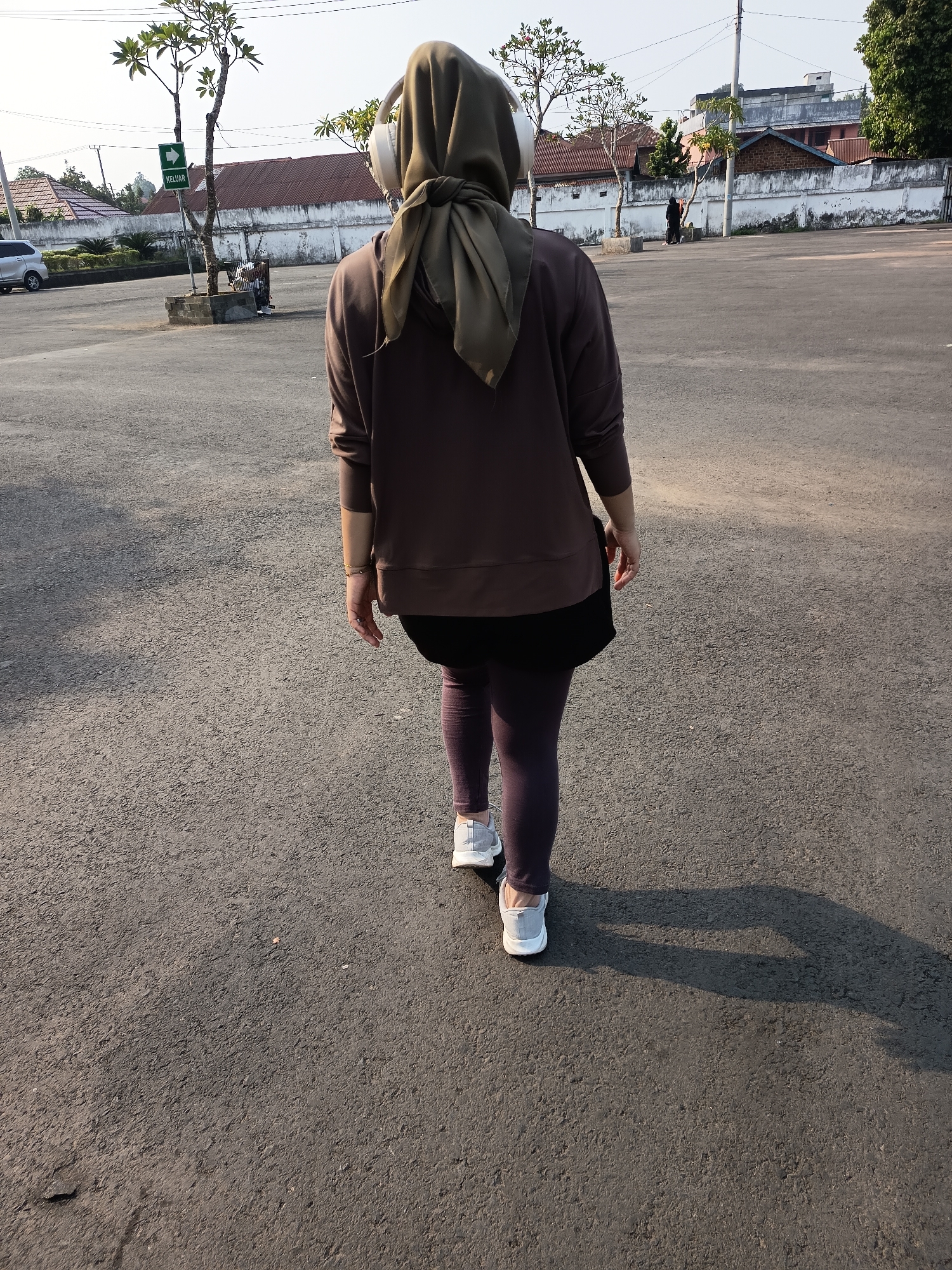 A person wearing a brown long-sleeved shirt, black shorts, and purple leggings walks away from the camera on an asphalt surface. They have a green hijab and white headphones on. The person is in Lubuklinggau, Indonesia, as indicated by a green sign with "KELUAR" (Indonesian for "Exit") and an arrow pointing left, which is visible in the background. The environment is an outdoor, paved area with some trees and buildings in the distance. The sky is clear and bright, suggesting daytime.