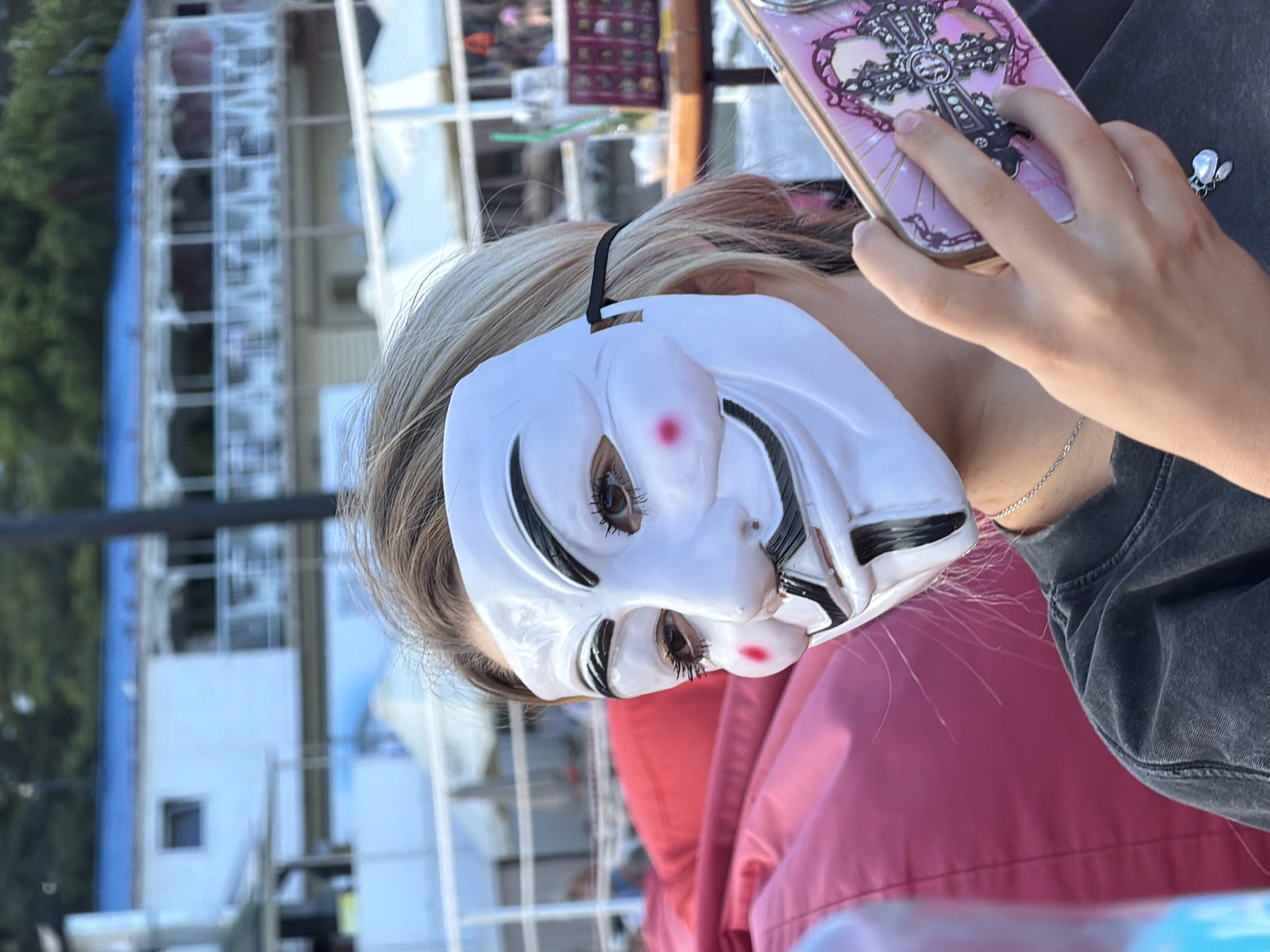 In Alushta, Ukraine, a person wearing a Guy Fawkes mask is partially visible, holding a pink phone with a cross design. They are seated in front of a large pink beanbag chair. In the background, a white building with a blue roof stands behind a railing. A wooden table with drinks and what appears to be a display of items is also visible. The scene suggests an outdoor setting, possibly a cafe or recreational area.