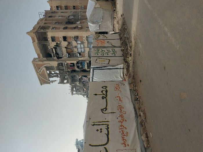 A partially destroyed building stands tall, with visible damage from what appears to be shelling or bombardment. Banners with Arabic script are displayed along the roadside. The surrounding area shows signs of conflict or destruction. The building's structure is severely compromised, exhibiting gaping holes and exposed interiors. A road and some temporary structures are also visible.