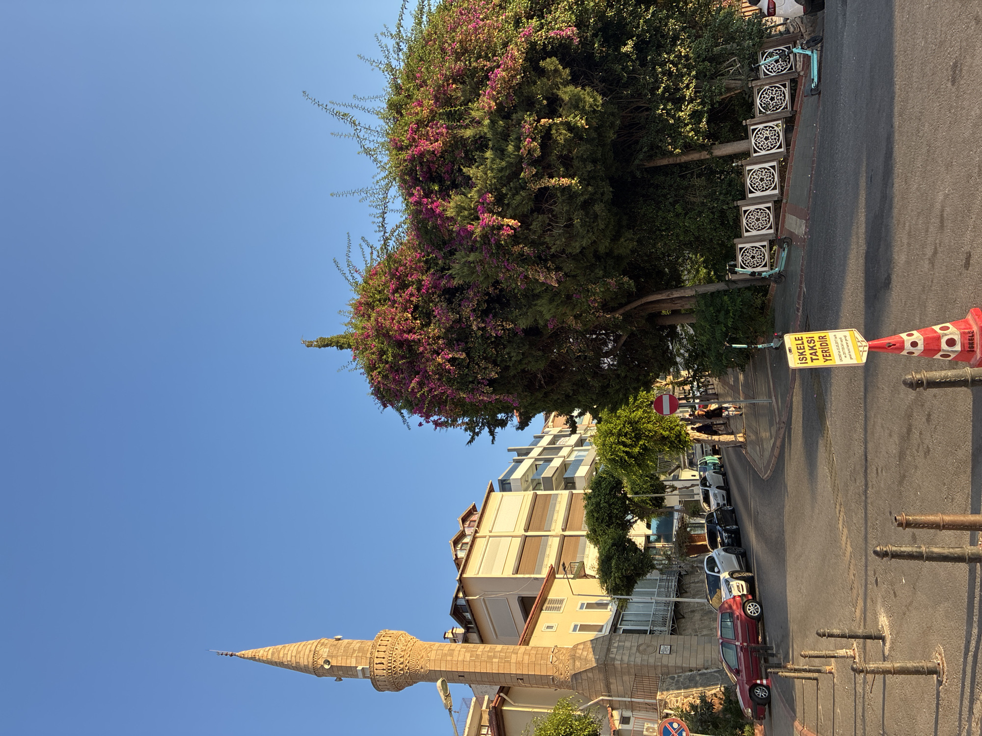 The image shows a street scene in Alanya, Turkey, with a prominent minaret on the left and a large, overgrown tree with pink and purple flowers on the right. Several buildings, including apartment complexes, line the street. Cars are parked along the side of the road. In the foreground, a yellow sign with Turkish text, placed on a traffic cone, indicates a taxi stand. Several electric scooters are visible parked near a decorative fence. The sky is clear and blue, suggesting it is a sunny day. The overall impression is a lively urban environment with hints of Mediterranean charm.