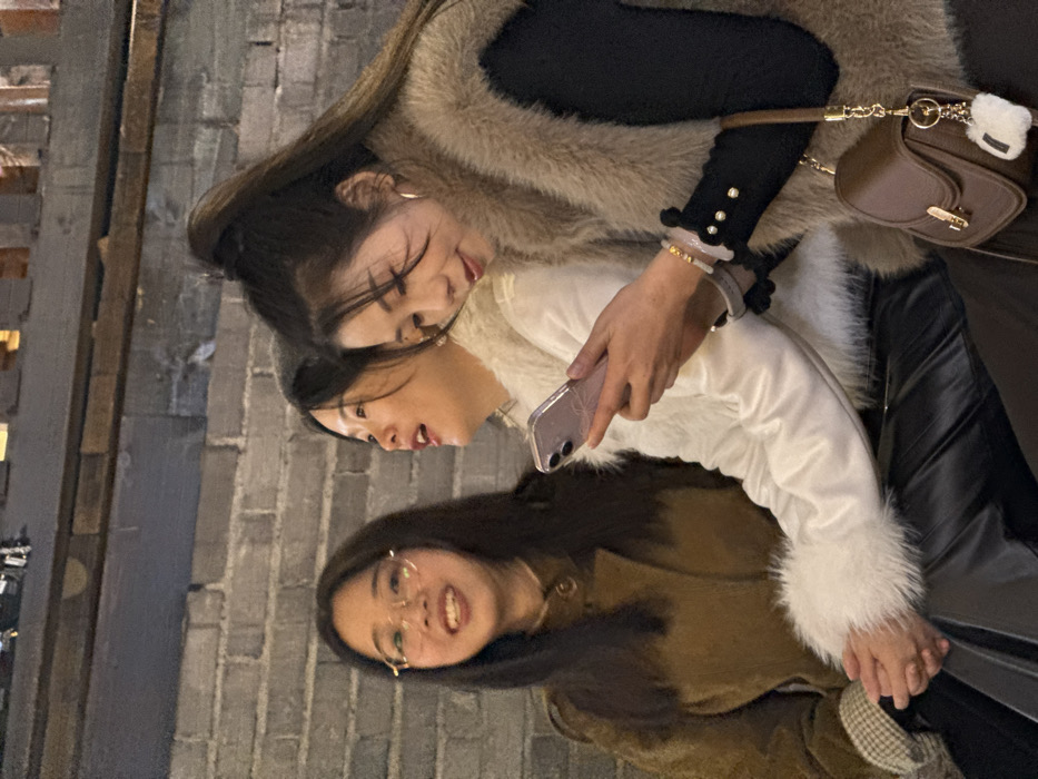 Three East Asian females are seated against a grey brick wall in Yuzhong District, China. The leftmost female, with long dark hair and gold-rimmed glasses, wears a brown button-up coat with a patterned inner cuff visible. She is smiling and looking towards the viewer's left. The central female, with dark hair, wears a white jacket with white faux fur trim on the collar and cuffs, paired with black pants. Her mouth is slightly open, and she is also looking towards the left. The rightmost female, with long dark hair pulled into a high ponytail, wears a tan faux fur vest over a black long-sleeved top with small pearl-like buttons on the cuff. She is holding a light purple smartphone with a patterned case in both hands, looking down at its screen. A small brown leather shoulder bag with a gold clasp and a white fluffy charm hangs on her left side. Multiple bracelets, including white and gold beads, are visible on her left wrist. The background features horizontally laid grey bricks, with a dark wooden beam or structure visible above them. The scene is illuminated by artificial light.