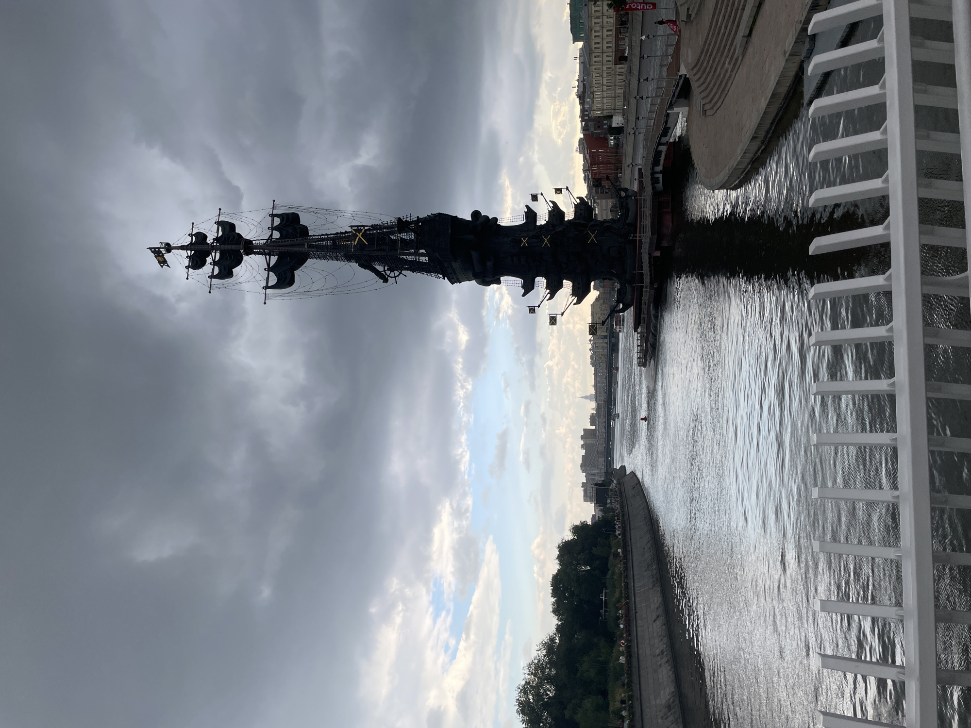 An outdoor, medium shot photograph depicts a large, dark-colored statue of a ship with a prominent mast and several decks, situated on the water. The statue is positioned on the right side of the frame, with its reflection visible in the rippling water. In the background, a city skyline with buildings of varying heights and architectural styles can be seen. A river flows from the left foreground towards the background, with a tree-lined embankment on the left side of the river. Several people are visible on the embankment, appearing to be walking or gathered. A white railing runs across the bottom of the frame, suggesting the photo was taken from a bridge or elevated walkway. The sky is heavily overcast with dark, dramatic clouds. The overall lighting suggests it is either early morning or late afternoon under cloudy conditions.
