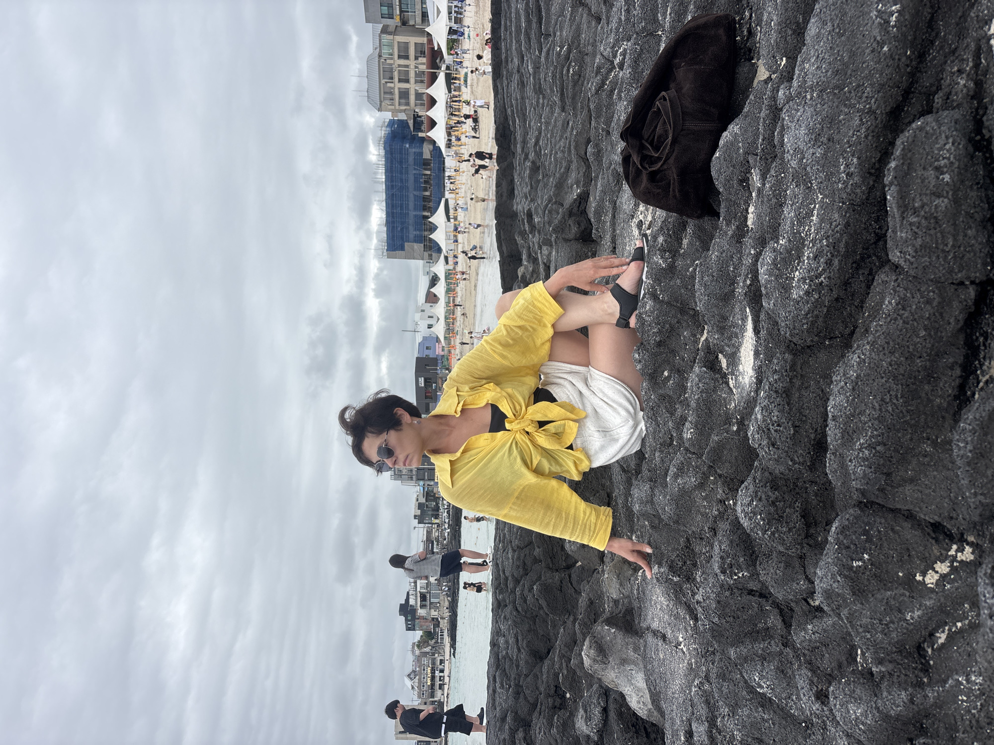 The image depicts a woman sitting on dark volcanic rocks by the sea, with a beach and buildings in the background. She is wearing a yellow, unbuttoned shirt tied at the waist over a black bikini top and white shorts. She also wears black sandals and sunglasses, with her dark hair styled in a messy bun. Her posture suggests a relaxed, casual pose, with one hand resting on the rocks and the other on her leg.
The setting is a beach with many people scattered across the sand and near the water. Tents and temporary structures are set up along the shoreline, indicating a possible event or a popular tourist spot. The buildings in the background are a mix of modern structures and older architecture, with scaffolding visible on one building, suggesting ongoing construction. The sky is overcast, indicating a cloudy day. The overall mood appears calm and leisurely, with the focus on the woman enjoying the seaside atmosphere. The location is identified as Jeju, South Korea. No visible text is present in the image.