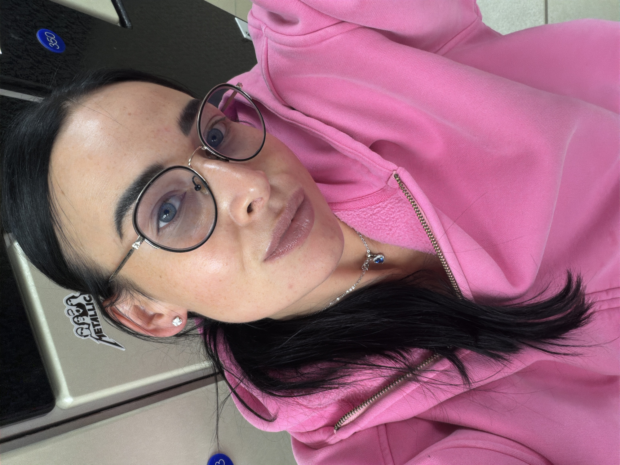 A close-up selfie captured indoors features a person, likely female, looking directly into the camera. The subject has long, dark hair, striking blue eyes, and wears round eyeglasses with thin, possibly metallic, frames. A light pink hooded sweatshirt (hoodie) covers her upper body, with a zipper visible down the front. A silver necklace with a small, blue-ish charm dangles just below her neck. A single diamond stud earring is visible in her left ear.
The background is somewhat indistinct but appears to be an indoor setting, possibly a locker room or a casual facility, suggested by beige/grey partitions or furniture. A black surface is also visible behind her head. To the left of her ear, a black and white sticker is affixed to the background surface. The sticker features the band name "METALLICA" in a jagged font, accompanied by stylized black figures. Above her head, a blue circular tag with the number "350" is partially visible on the black background.
The lighting is consistent with indoor conditions, and the person's expression is neutral, with slightly pursed lips. The image conveys a casual moment. The location is contextualized as Saint Petersburg, Russia.