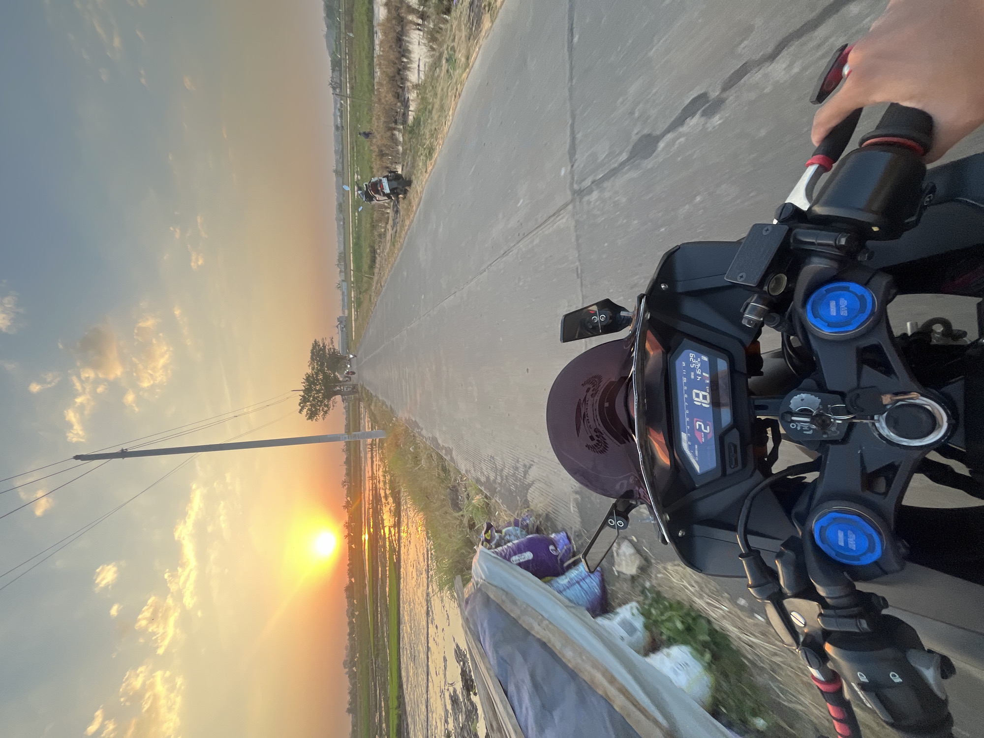 The image captures a first-person perspective of riding a motorcycle down a paved road in Pecangaan, Indonesia, during sunset. The sun, a bright orb low in the sky, casts a warm golden and orange glow across the scene, illuminating scattered clouds. To the left of the road, a flooded rice field reflects the sky's colors, bordered by green grass and some debris on the edge of the road. To the right, a person is visible standing near a parked motorcycle in a grassy field. The road stretches forward, leading towards a distant line of trees and structures, with a utility pole standing prominently on the left side. The motorcycle's handlebars, dashboard displaying speed and time, and a hand on the throttle are in the foreground, indicating the motion of the ride.