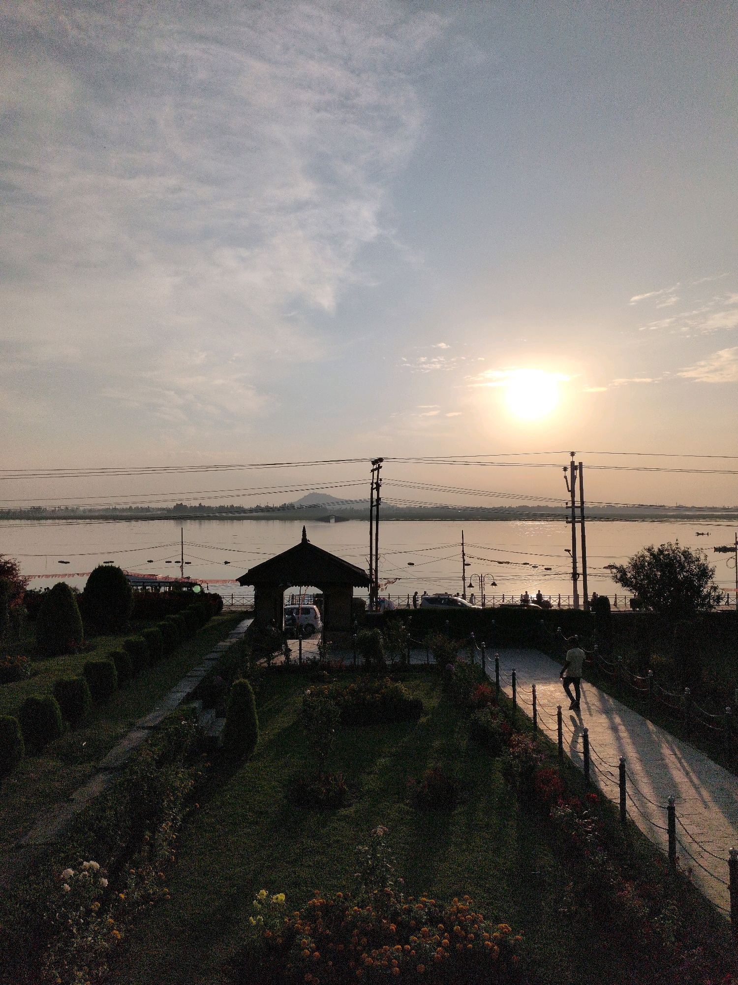 The image captures a serene sunset scene at the Dal Lake in Srinagar, India. The sun is low on the horizon, casting a warm, golden glow across the water and sky, with a few wispy clouds dotting the upper portion of the frame. In the foreground, a well-maintained garden is visible, with manicured hedges and blooming flowers. A paved pathway winds through the garden, bordered by a decorative fence. A lone figure walks along the path, their silhouette cast by the setting sun. Further back, a small, open-air pavilion with a traditional roof structure stands near the water's edge. Across the lake, the distant shoreline is lined with trees and hills, bathed in the soft light of dusk. Several boats are scattered on the lake, adding to the picturesque landscape. Utility poles and power lines are also present in the scene, spanning across the middle ground.