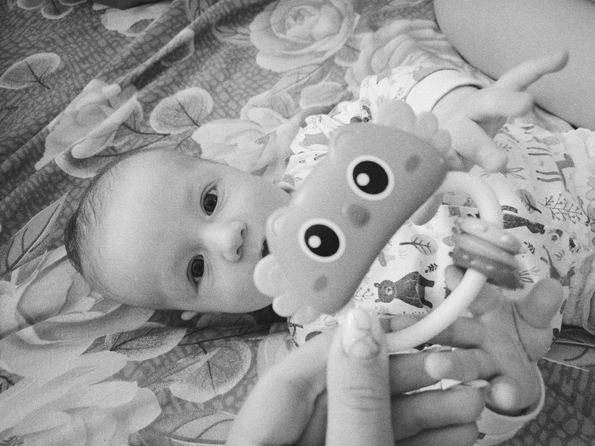 A black and white photograph shows a baby lying on a floral patterned blanket or sheet. The baby is looking towards the camera. A toy, possibly made of plastic and with large circular eyes, is positioned between the baby and the camera. The baby's hands are holding the toy. The scene is indoors. There is no indication of this photo being taken in Tobolsk, Russia.
