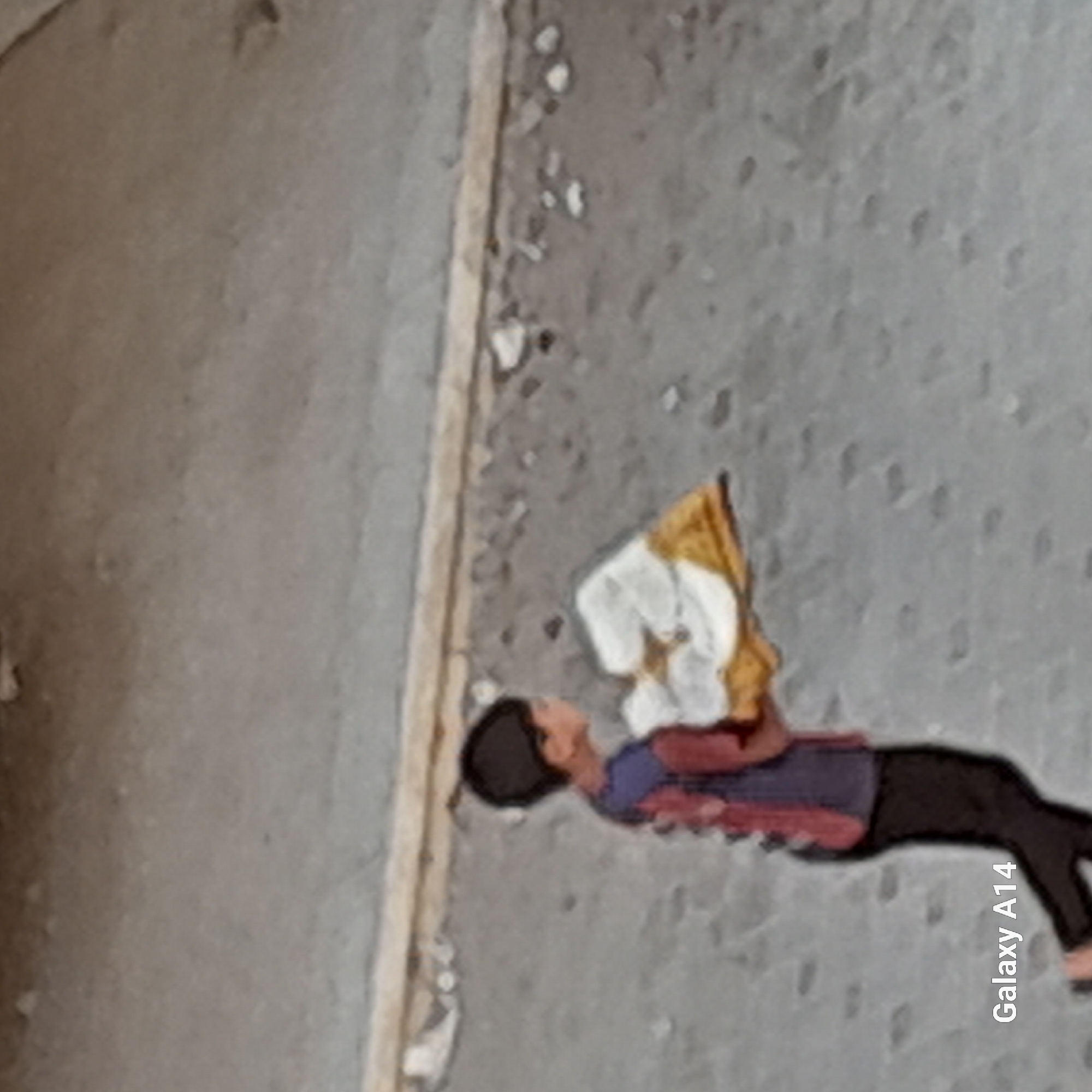 A young boy walks across a dusty, grey surface in Khan Yunis, Palestinian Territories, carrying a folded piece of yellow and white fabric. The boy is wearing dark pants and a shirt with red and blue accents. The background is a textured, concrete-like surface with scattered debris and a horizontal band of lighter material. The image is low resolution and has a stylized, almost painted appearance. The text "Galaxy A14" is visible in the bottom left corner, indicating the device used to capture the image.