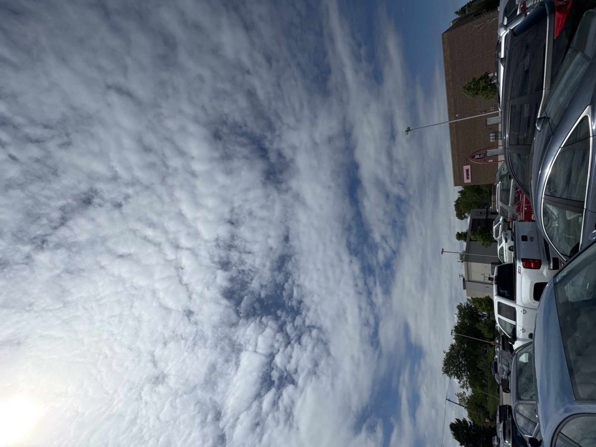 The image shows a parking lot in Glendale Heights, United States of America. The sky is mostly covered with white, puffy clouds, with patches of blue visible in between. Several parked cars are visible, some of which are silver or gray. A building, possibly a retail store, is visible in the background. Trees are also visible along the horizon line. A street light is visible near the building.