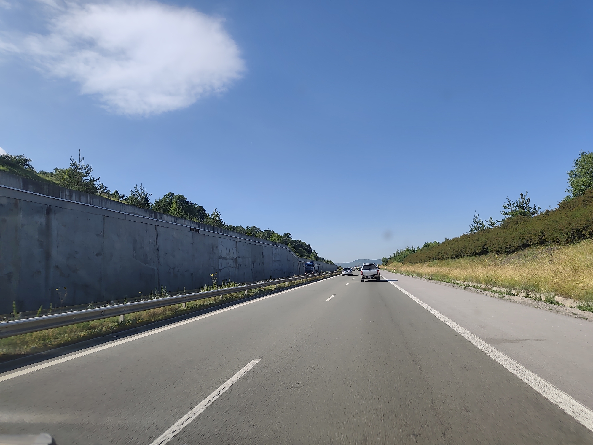 The image shows a bright, sunny day on a highway in Golemo Buchino, Bulgaria. The perspective is from inside a vehicle traveling on the road. To the left, there is a concrete retaining wall that slopes upwards, topped with green trees and vegetation. A metal guardrail runs along the edge of the road in front of the wall. The highway itself is paved with asphalt and has white lane markings. In the distance, other vehicles can be seen, including a dark truck, a white car, and a silver SUV. The right side of the highway features a grassy embankment covered with dry yellow grass and more green trees. The sky is a clear, vibrant blue with a large, fluffy white cloud in the upper left quadrant. The overall scene depicts a typical highway environment in a rural or semi-rural setting.