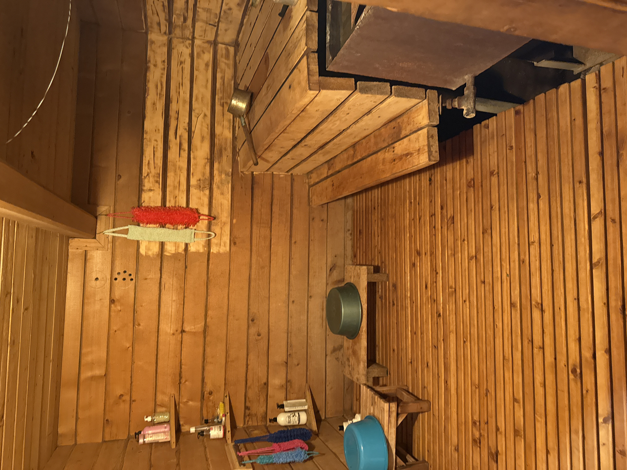 The image depicts the interior of a wooden sauna. The walls and ceiling are constructed from horizontal wooden planks, giving the space a rustic ambiance. In the foreground, a wooden floor made of narrow planks stretches across the room. To the left, there are two low wooden benches, each holding a plastic basin; one is blue, and the other is a greenish-gray. Above these benches, a wooden shelf holds several bottles of toiletries. A string of colorful bath brushes hangs on the left wall, near the toiletries.
In the midground, a multi-tiered wooden bench system dominates the right side of the sauna, designed for seating. On the top level of this bench, a metal ladle rests on the wooden planks. To the right of the benches, a large, dark metal container, possibly a heater or water tank, is partially visible. The overall lighting is warm and subdued, characteristic of an indoor sauna environment. The location is identified as a sauna in Чуй-Атасево, Russia. There are no people visible in the image, and no discernable actions or interactions are taking place.